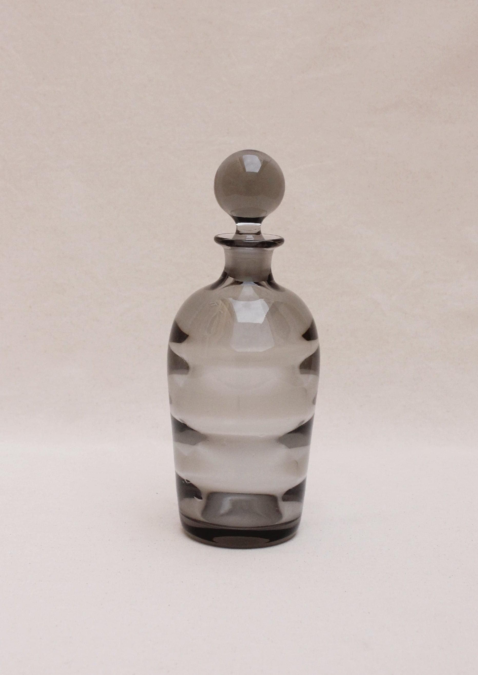 An Boga Avante Shop Art Deco liquor decanter, transparent with a rounded stopper, features alternating clear and frosted horizontal bands in an optical glass design, set against a plain, light beige background.
