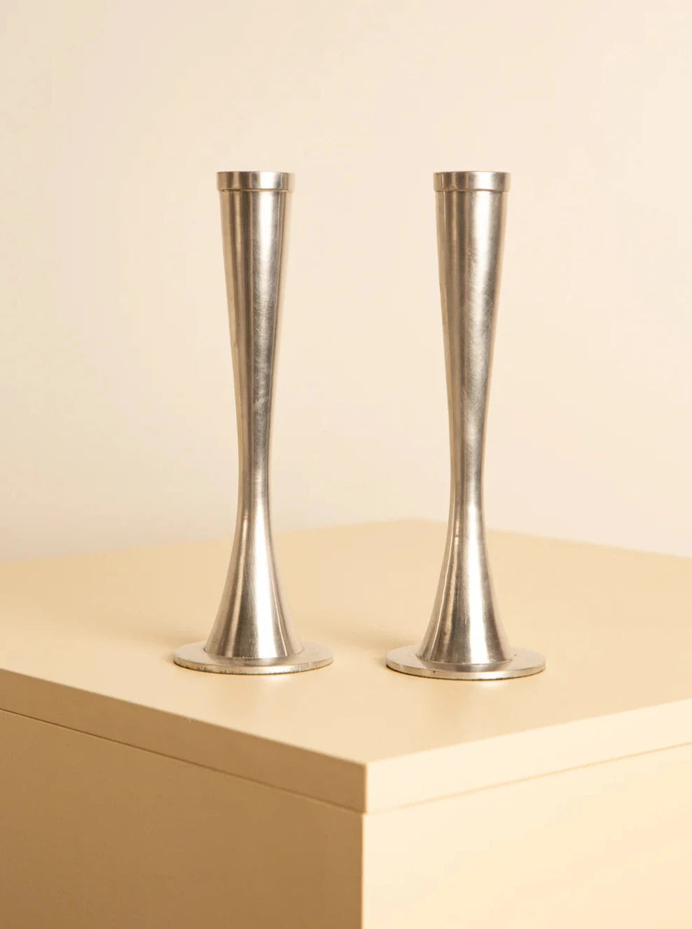 Two sleek Treaptyque Pair of 80's aluminum candleholders stand side by side on a light-colored surface. Their design is simple and elegant, with a narrow mid-section and a slightly flared base and top. The background is plain and beige, highlighting the minimalist aesthetic and showcasing meticulous craftsmanship.