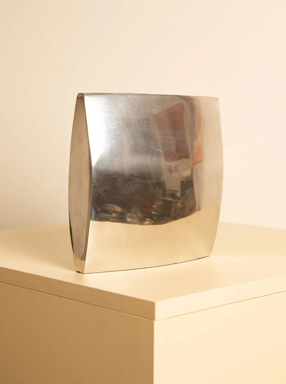 The 80's Italian metal vase by Treaptyque, with its curved metallic rectangular form and reflective surface, is elegantly perched on a beige pedestal. The backdrop is a plain light-colored wall, casting minimal shadows and reflections on the vase's metal surface, beautifully evoking the vintage elegance of Italian craftsmanship.