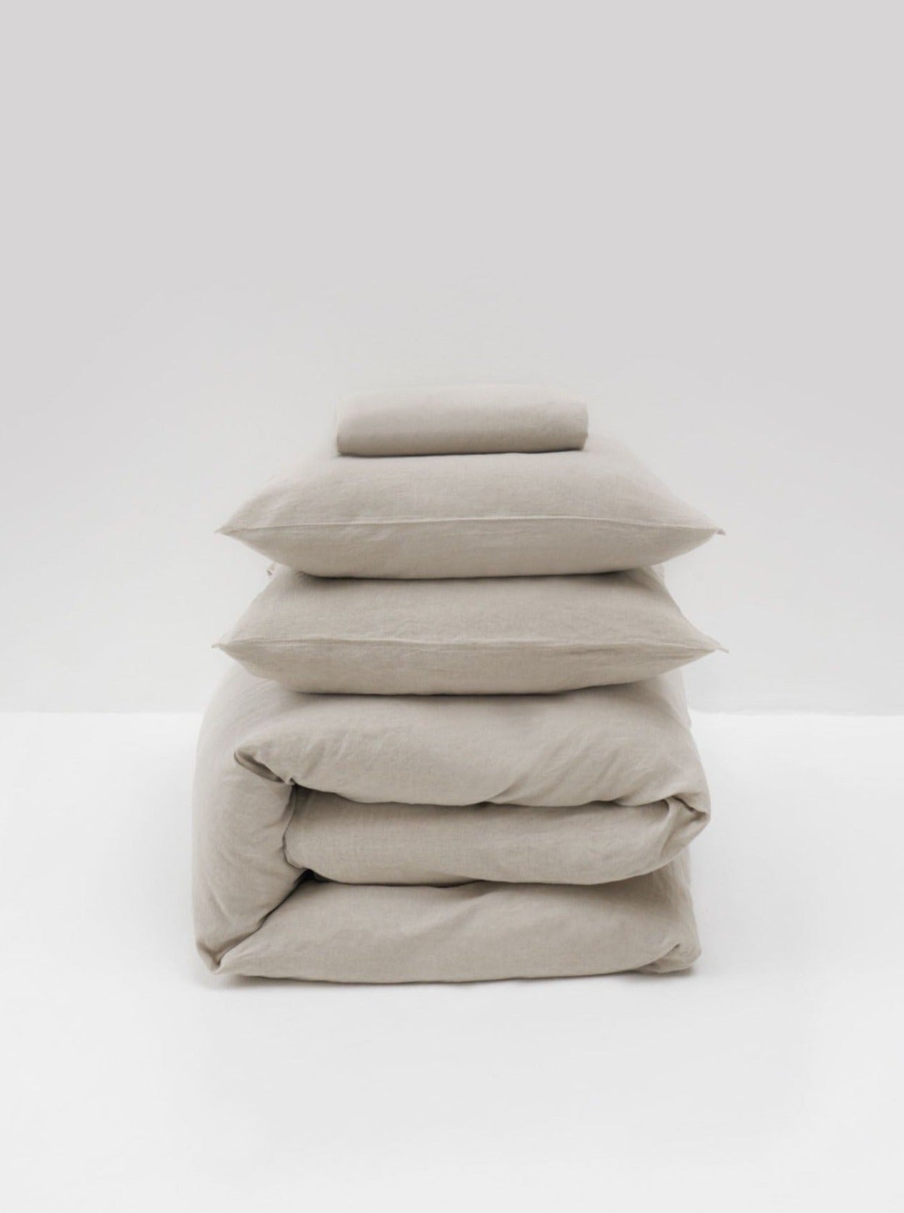 A minimalist display against a white background features the MOST Washed Linen Bed Set in Natural, showcasing neatly stacked beige bedding with two pillows and a folded duvet.