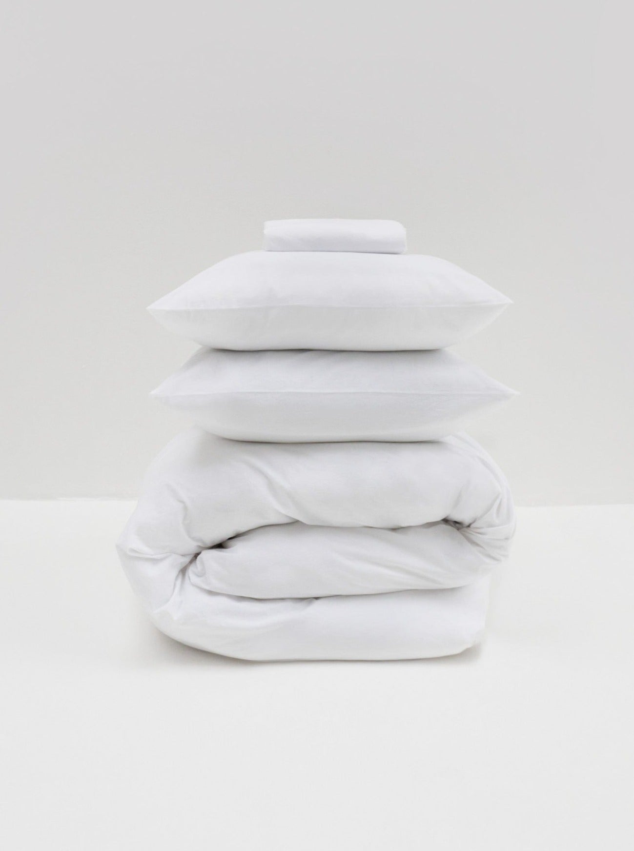 A neatly stacked set of MOST White Washed Cotton Percale Bed Set comprising pillows, a folded comforter cover, and sheets, all against a plain gray background.