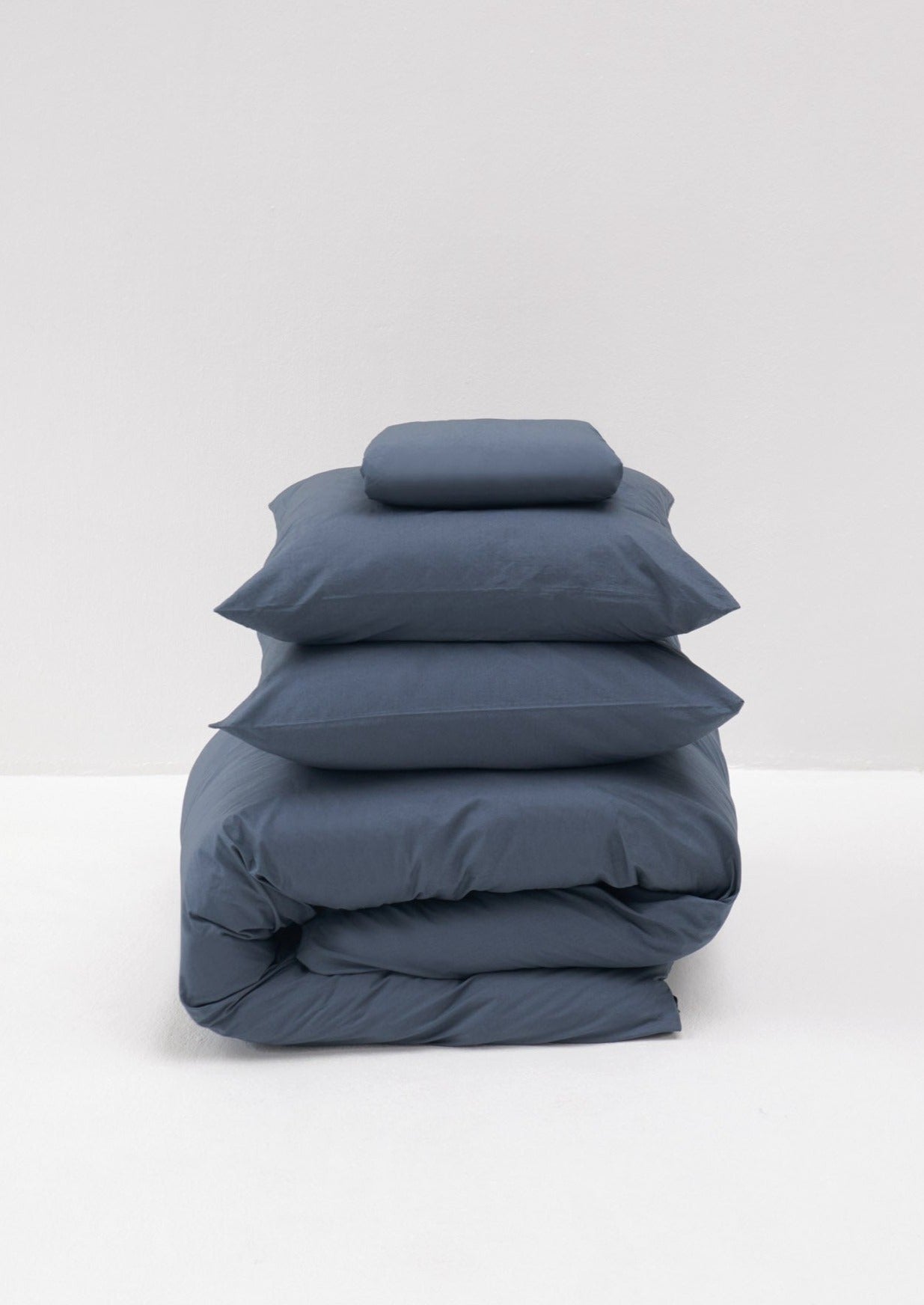 A stack of neatly folded MOST Cotton Percale in Stormy bed linen set, including a comforter cover and pillows, set against a simple, light gray background.