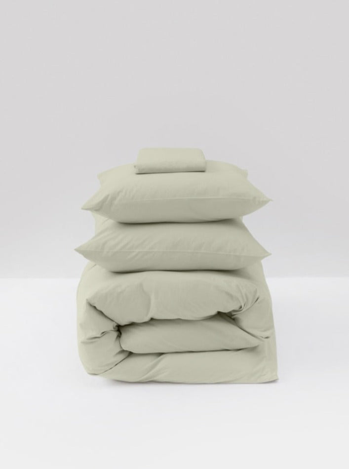 A neatly stacked set of MOST's Washed Cotton Percale Bed Set in Sage, including a folded comforter cover, sheets, and fluffed pillows, on a plain light background.