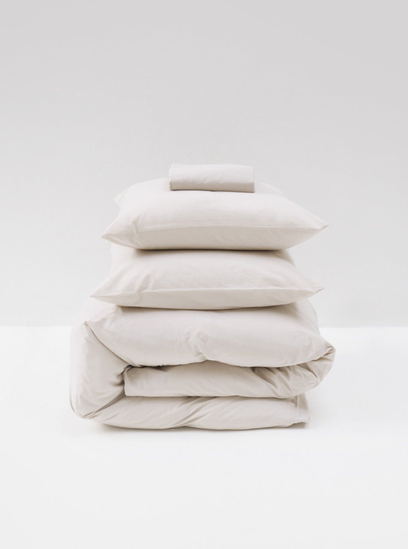 A neat stack of MOST's Washed Cotton Percale Bed Set in Sand, including two pillows, two duvets or comforter covers, and a folded towel, all in shades of white and cream, against a white background.