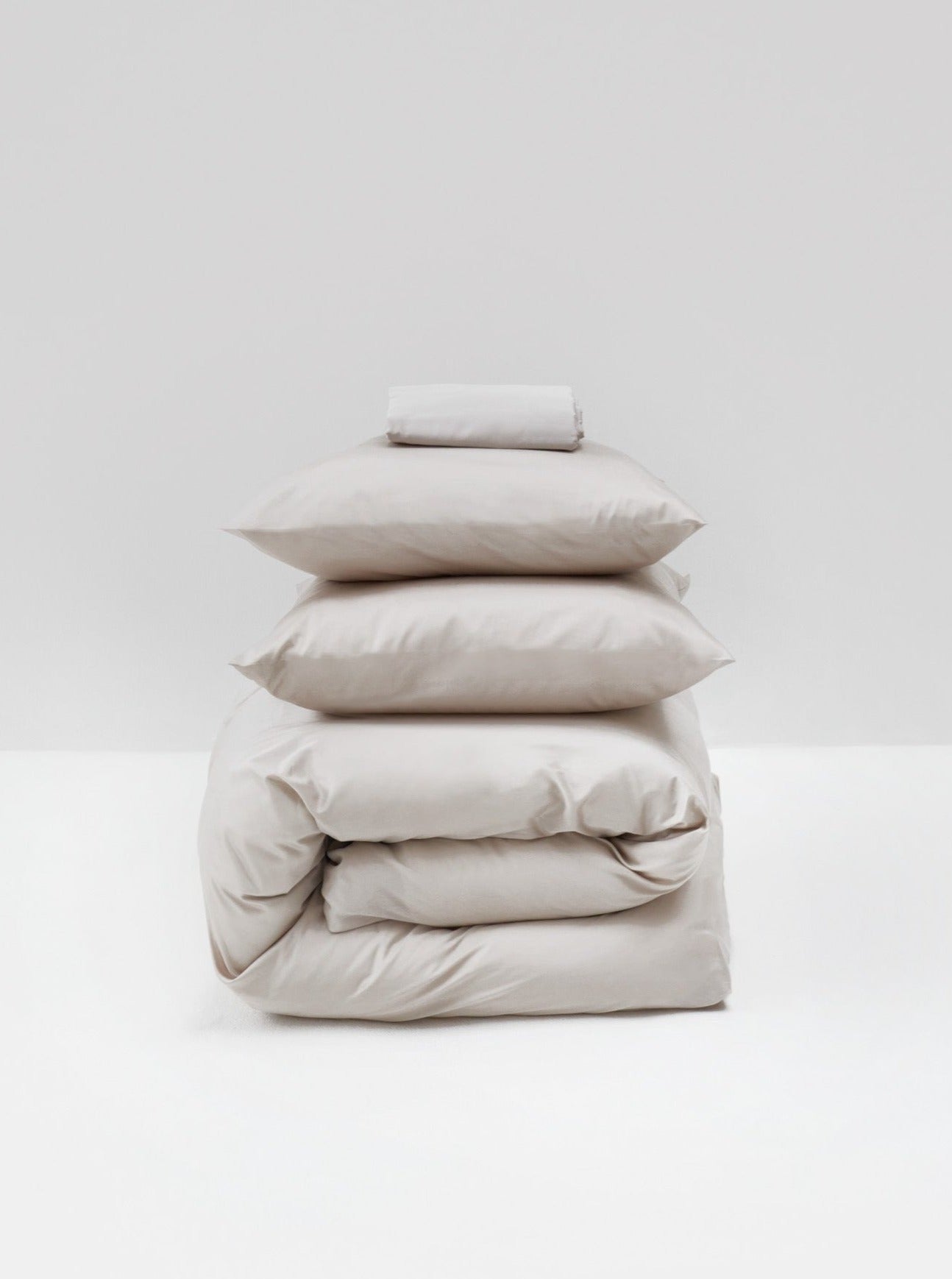 A stack of neatly folded MOST Cotton Sateen Bed Set in Sand bedding items including sheets, pillowcases, and a comforter cover against a plain white background.