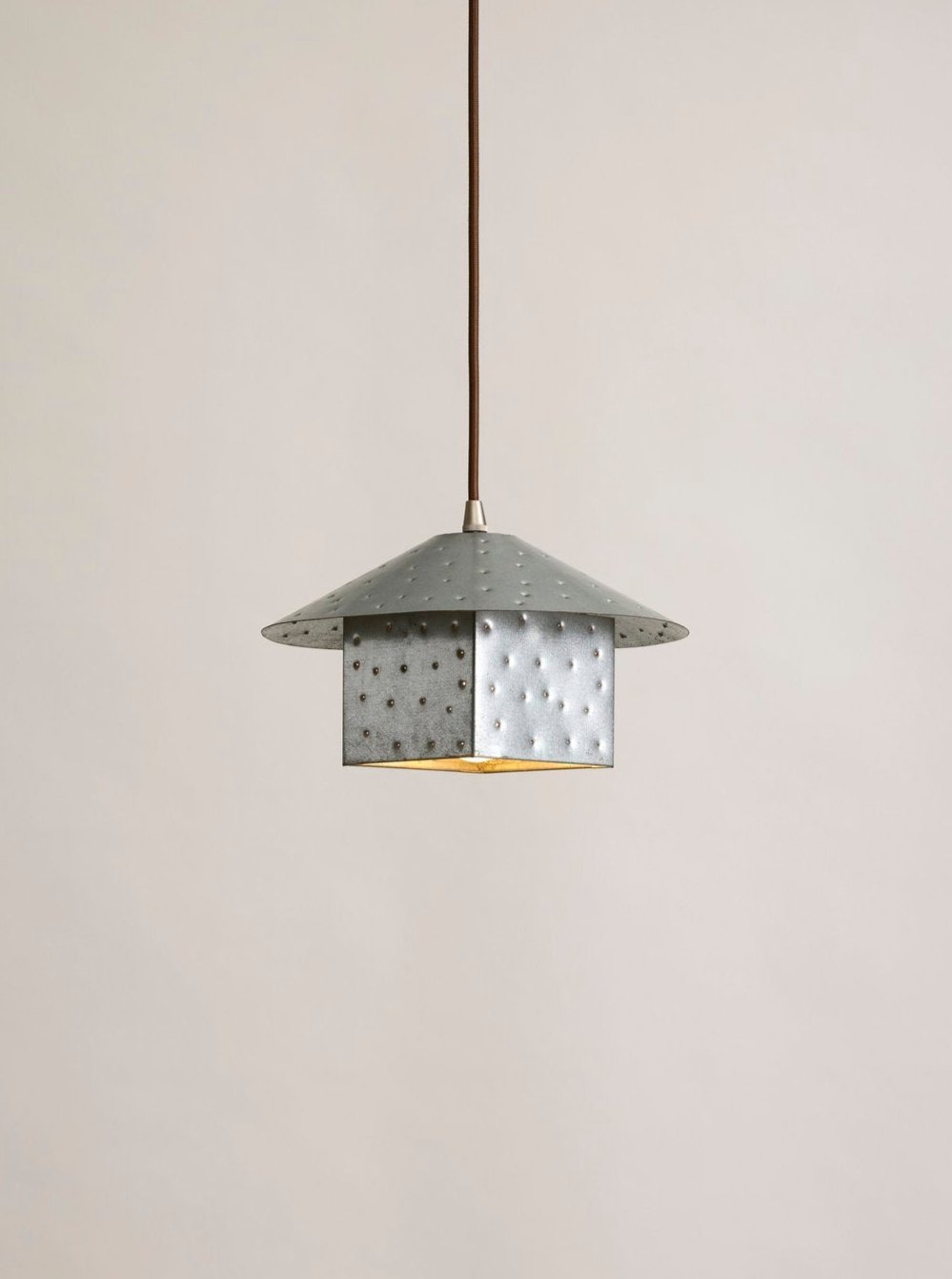 The Lin Pendant, Steel, Small by MUHLY is a modern light fixture inspired by environmental artist Maya Lin. It features a circular, flat shade in matte gray with hand-punched perforations and a central cube-shaped element that reflects her minimalist design. This elegant piece hangs from a single thin rod against a plain beige background.