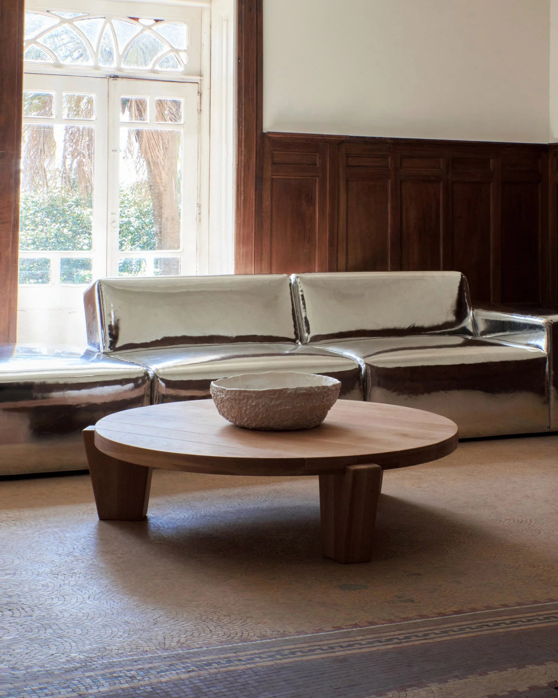 A modern living room showcases a metallic, reflective sectional sofa against wooden wall paneling. At the center, a "Peniche" Coffee Table in Oak by Project 213A, handmade in Portugal with solid oak construction, sits proudly adorned with a textured bowl. Natural light streams in through a window with a decorative archway.