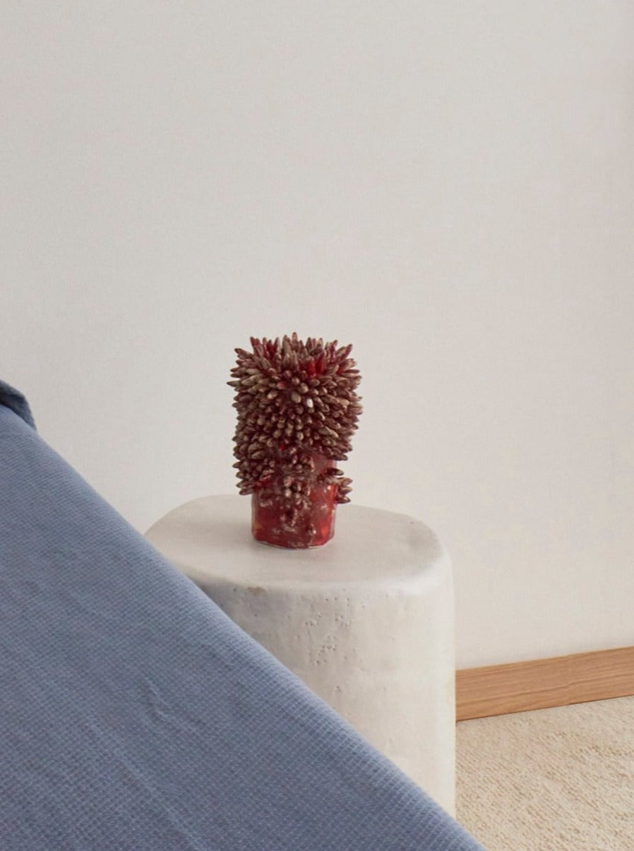 A small, red, textured handcrafted Appuntito Vase by Project 213A stands atop a white rectangular block beside a blue fabric edge, against a neutral-toned wall and floor backdrop.