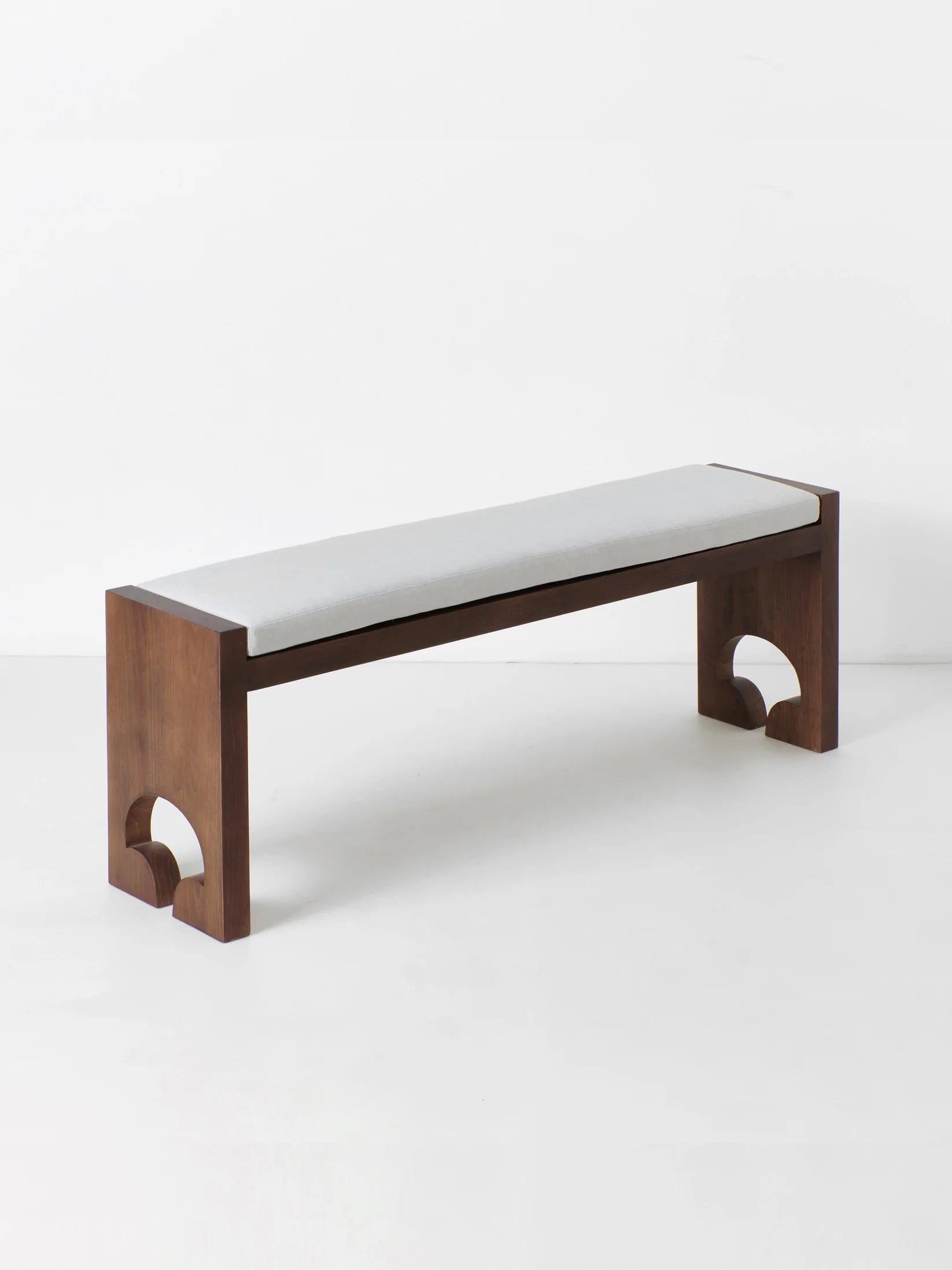 The Quinta Bench by Project 213A is a modern piece with a minimalist design, offering bespoke dimensions. Its rectangular seat is upholstered in light-gray fabric, and the unique cutout shapes on the thick, square legs add a touch of elegance while firmly supporting the solid wood structure.