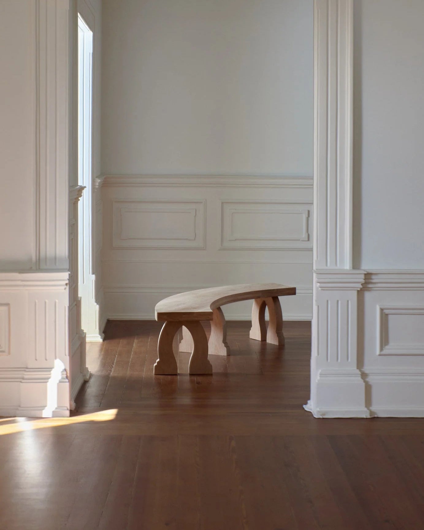A minimalist interior features white-paneled walls, a high ceiling, and a wooden floor. At the center of the room is a Project 213A "Sintra" Curved Bench in Oak, crafted by expert artisans. Sunlight streaming through a large window enhances the calm and serene atmosphere.