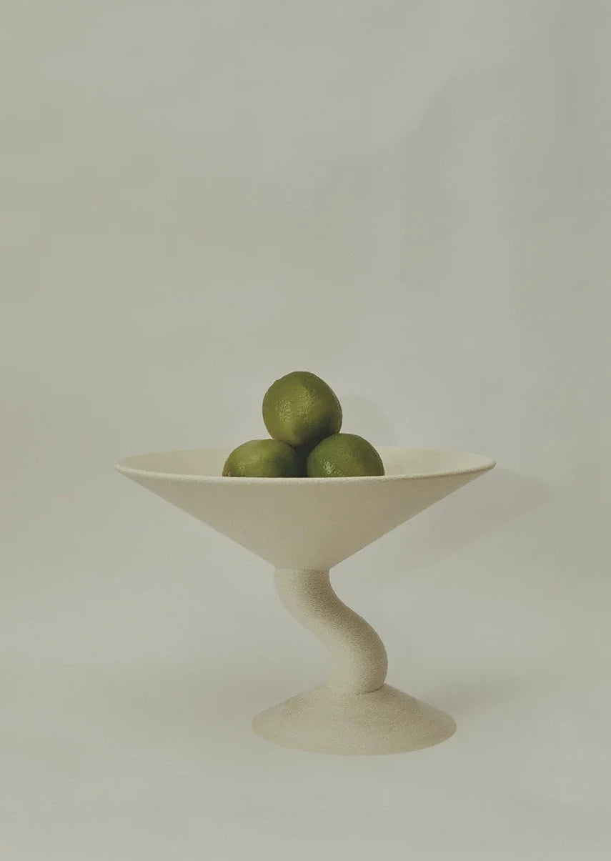 A minimalist image featuring a unique, sculptural Wavy Fruit Stand by Argot Studio, holding several small green limes, set against a plain, light background. This decorative bowl introduces an aesthetic blend of functionality.