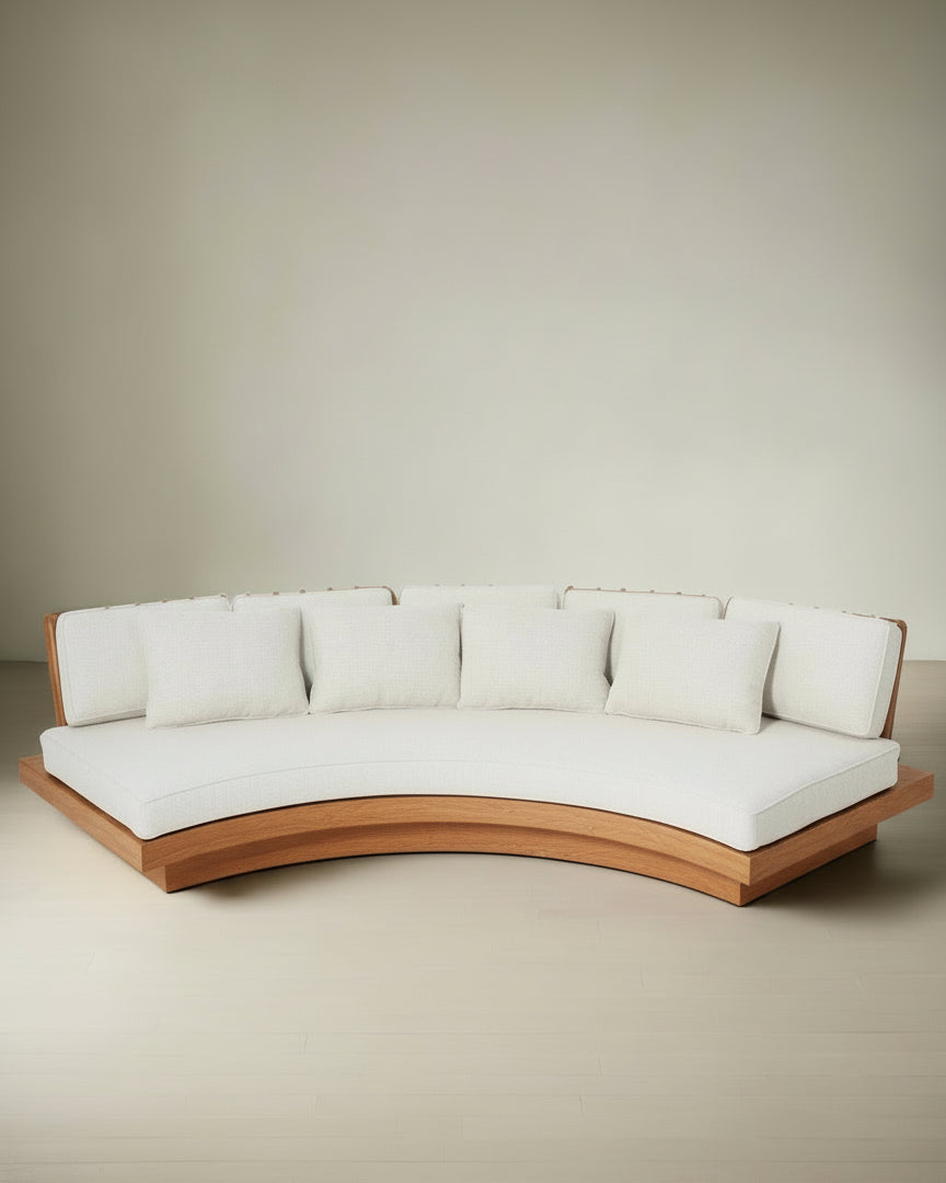 The Barracuda Interiors San Romano Round Oak Sofa, featuring a curved oak base and white cushions with five matching back pillows, sits elegantly against a light-colored wall and floor.