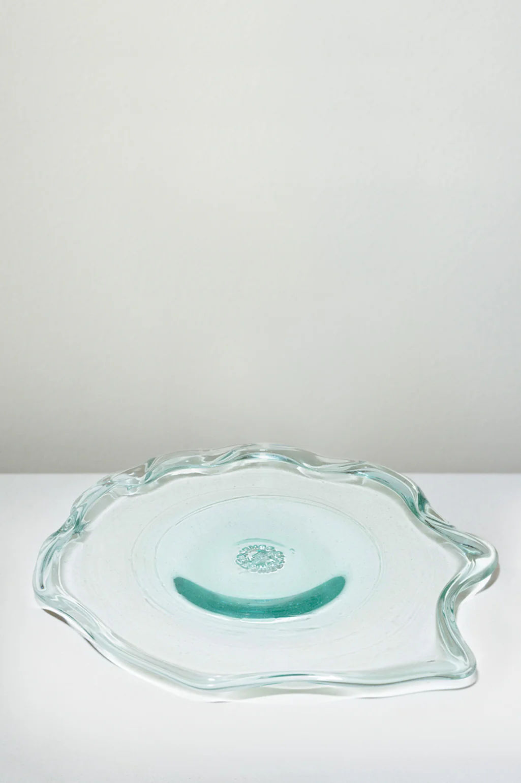 The Completedworks Pond serving plate is crafted from recycled glass with a clear, wavy-edged, irregular shape and subtle aqua tint, shown resting on a white surface against a light gray background.