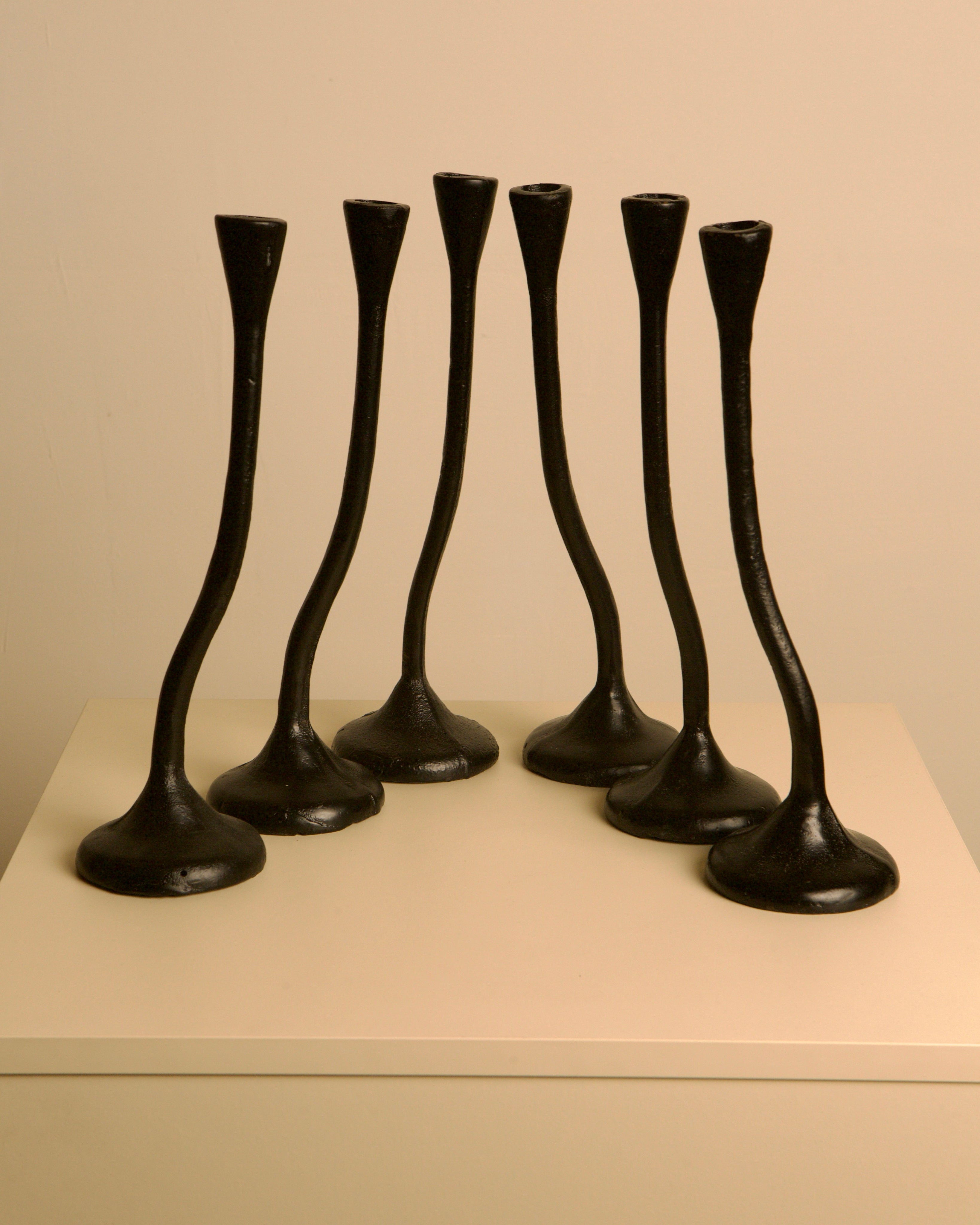 A set of 5 Treaptyque "Twisted" black cast aluminum candleholders from the 80s, featuring flared bases, is arranged on a light square surface against a plain beige background, showcasing sculptural design.