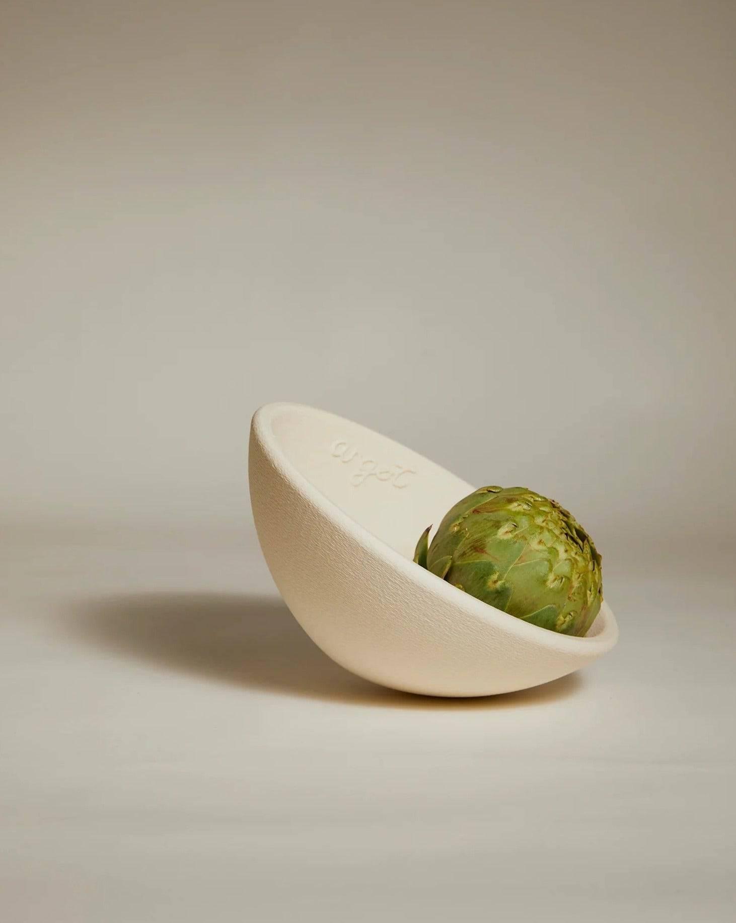 A green artichoke rests in the Acorn Bowl Grand by Argot Studio, a white ceramic bowl with a round bottom, slightly tilted against a plain beige background. The subtle texture and embossed text embody minimalism, enhancing its sculptural contemporary design.