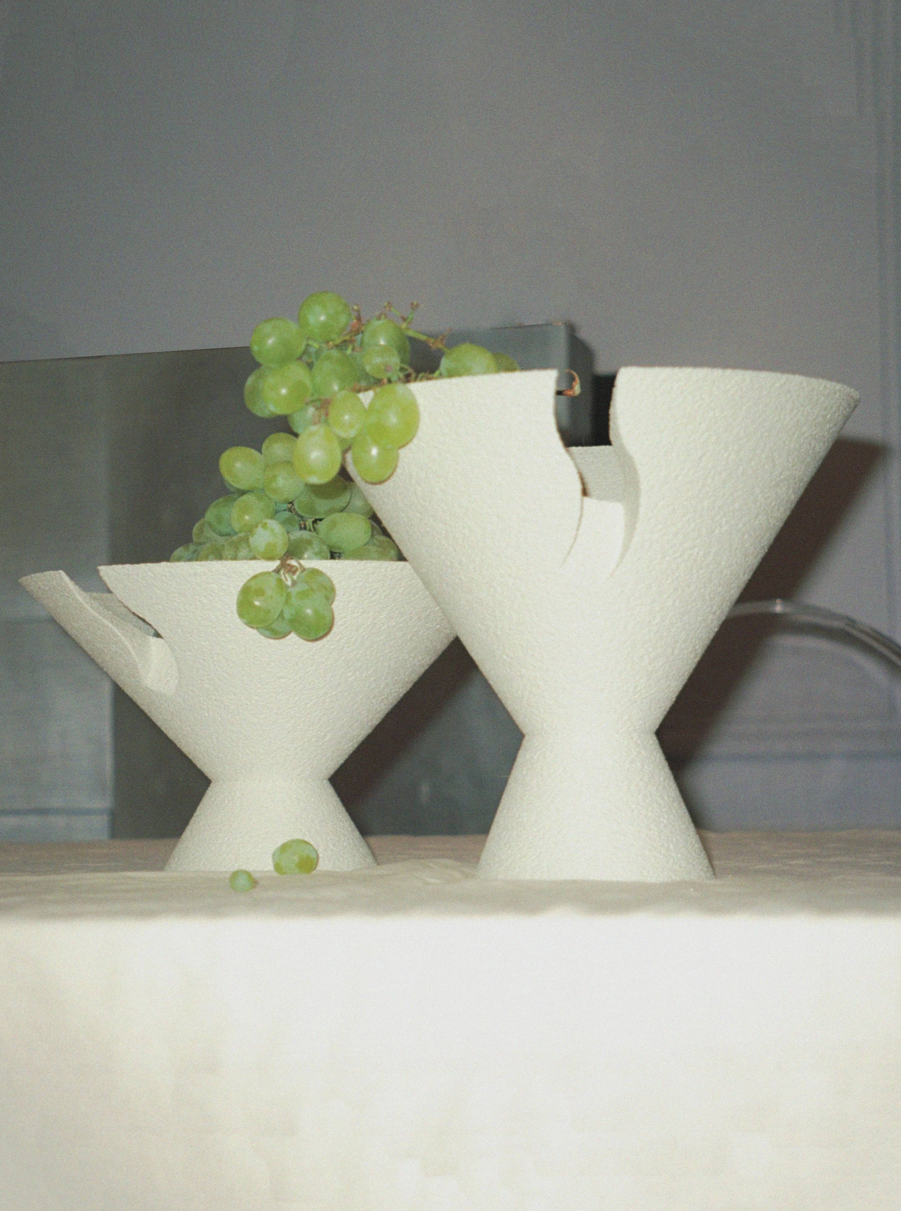 Two Tyro Bowls from Argot Studio, crafted using 3D printing technology with unique, abstract shapes, sit on a counter, one holding a bunch of green grapes. The background is softly out of focus.