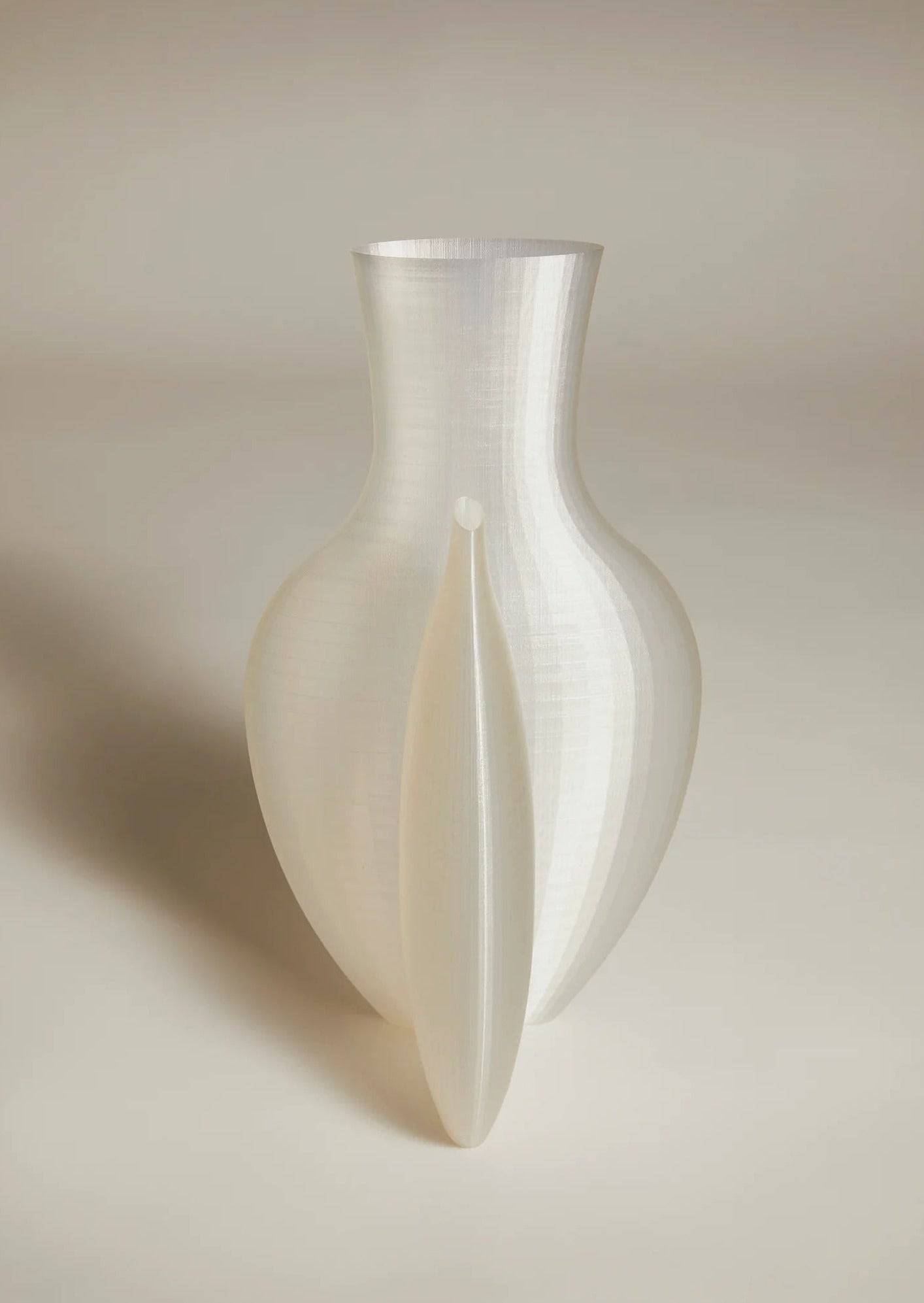 The Double Vessel - New Edition by Argot Studio is a translucent, abstract vase with smooth curves set against a neutral backdrop. This luxury piece boasts a vertical ripple pattern and an elongated, pointed detail, making it a unique collectible for any interior.