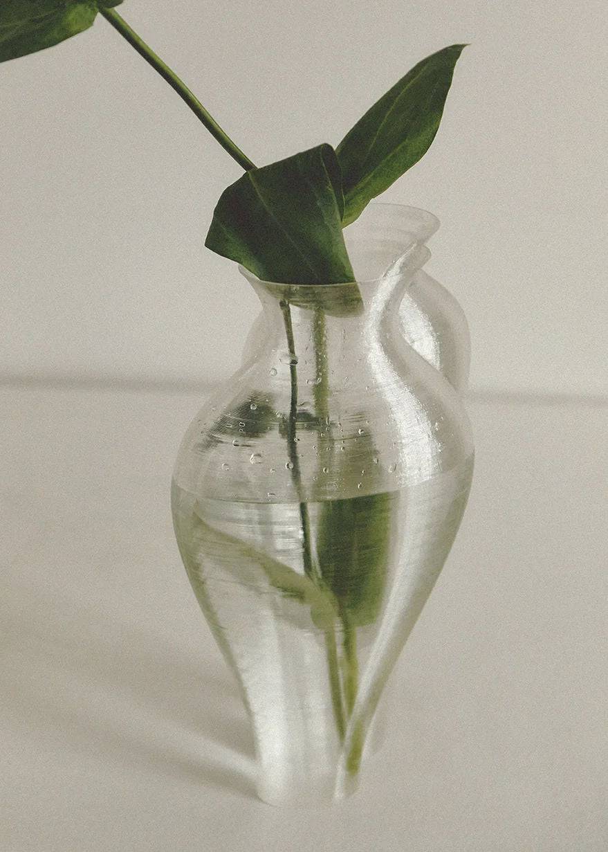 A Soeurs Vase by Argot Studio with a minimalistic design containing water and a single green leafy plant stem, placed on a neutral beige surface, casting a soft shadow.