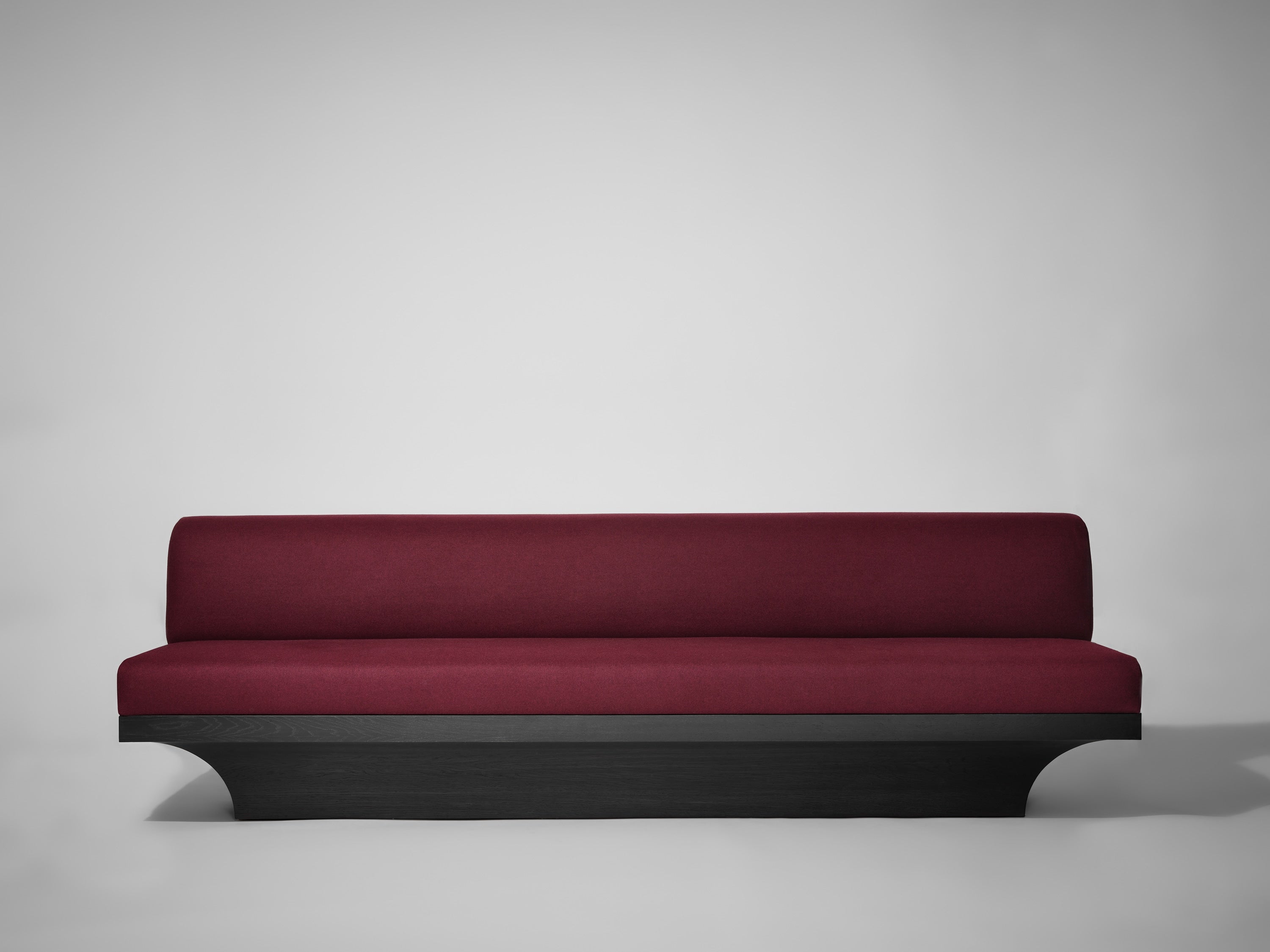 The Mokko Acer Sofa features a modern, minimalist design with a deep burgundy upholstered seat and backrest on a sleek black wooden base, making it an ideal statement piece for any living room.