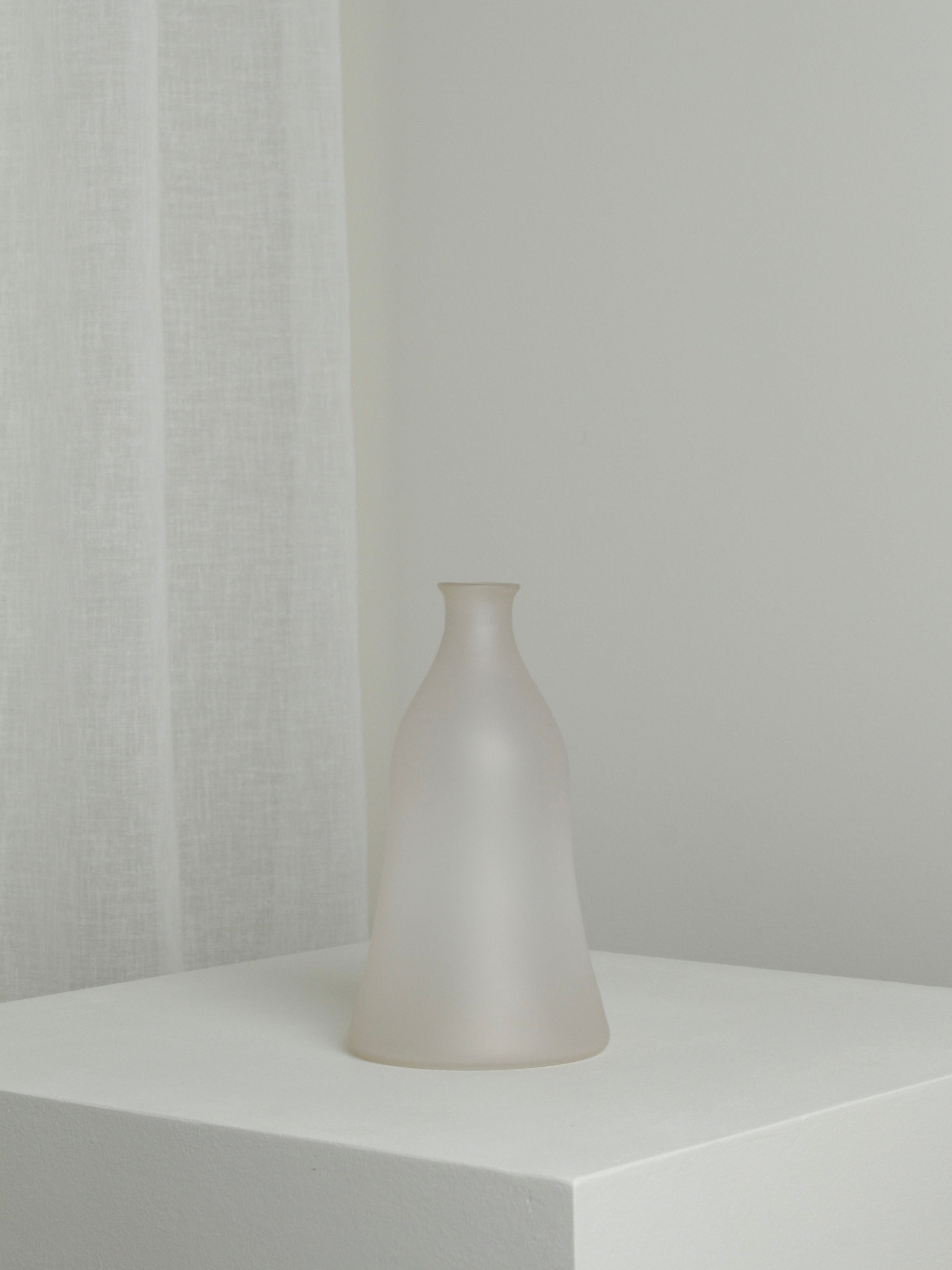 The Medium Vase in Moon by Verre d'Onge, featuring frosted glass and a narrow neck, is displayed on a white pedestal against a light gray wall and sheer curtain in a minimalistic, softly lit setting.