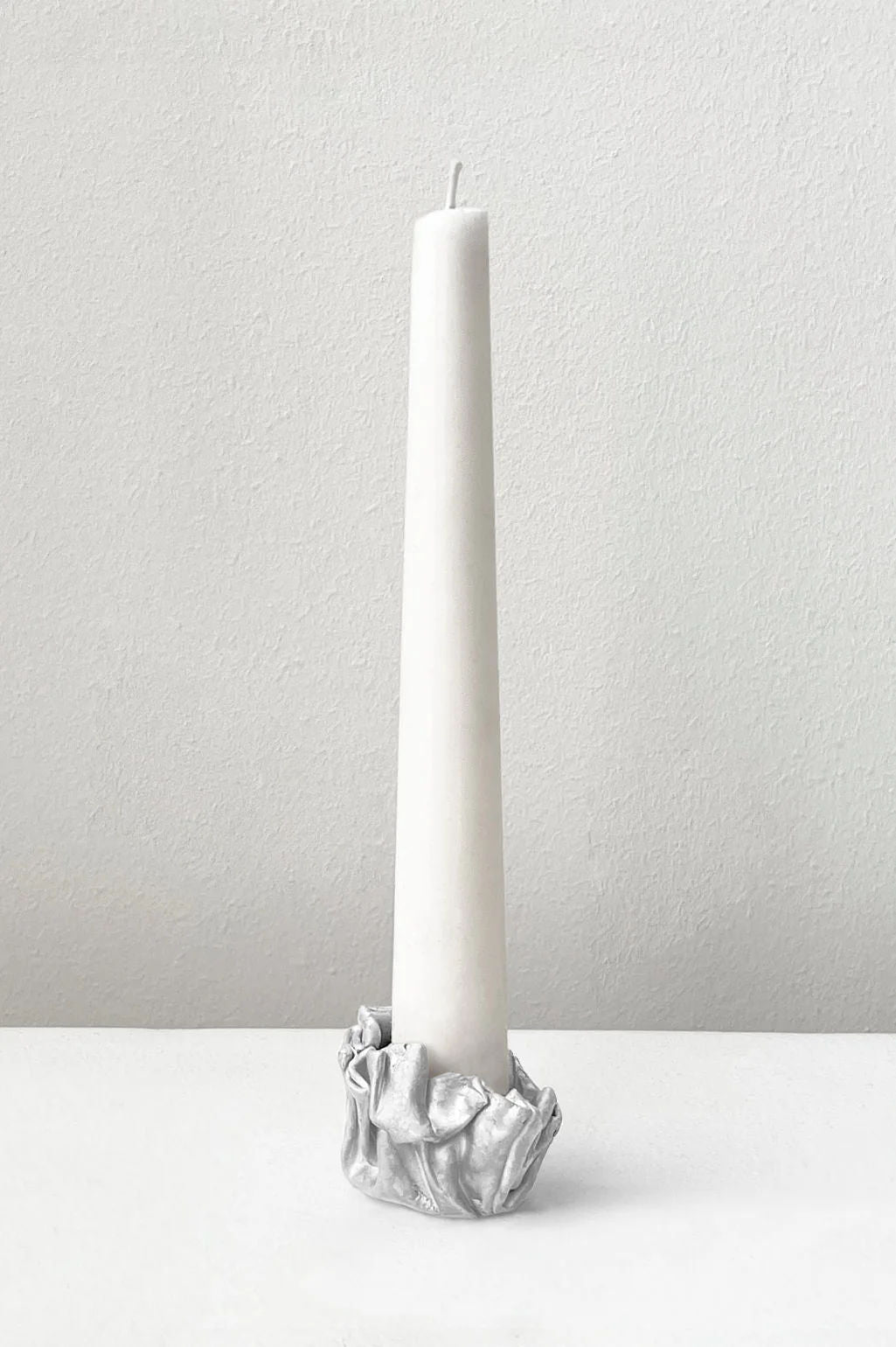 A tall white taper candle stands in the Completedworks L42 - Candle Holder in Silver, its metallic crumpled design shown against a plain white background.