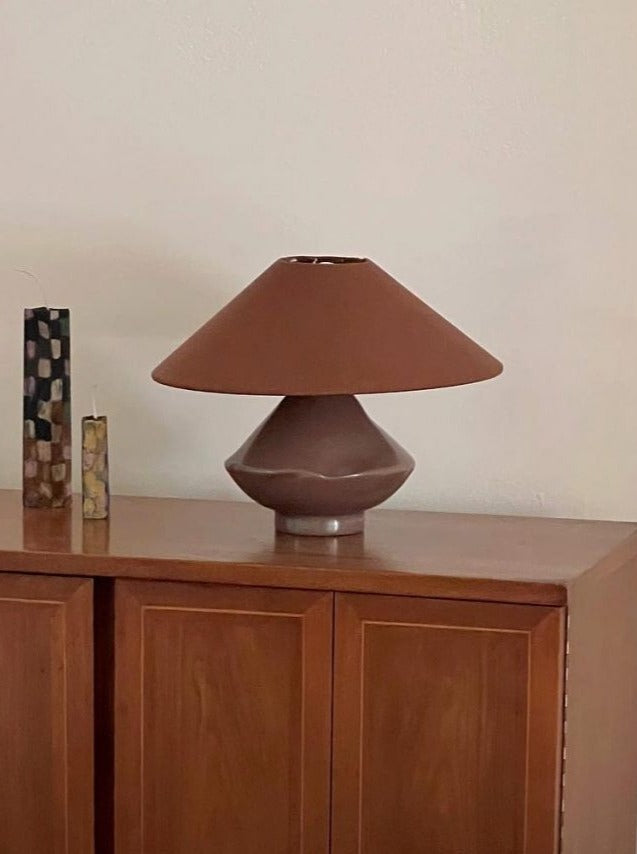 A mid-century modern-style Los Objetos Decorativos handcrafted lamp with a unique rounded triangular base and a matching shade, positioned on a wooden cabinet beside a decorative checkered vase.