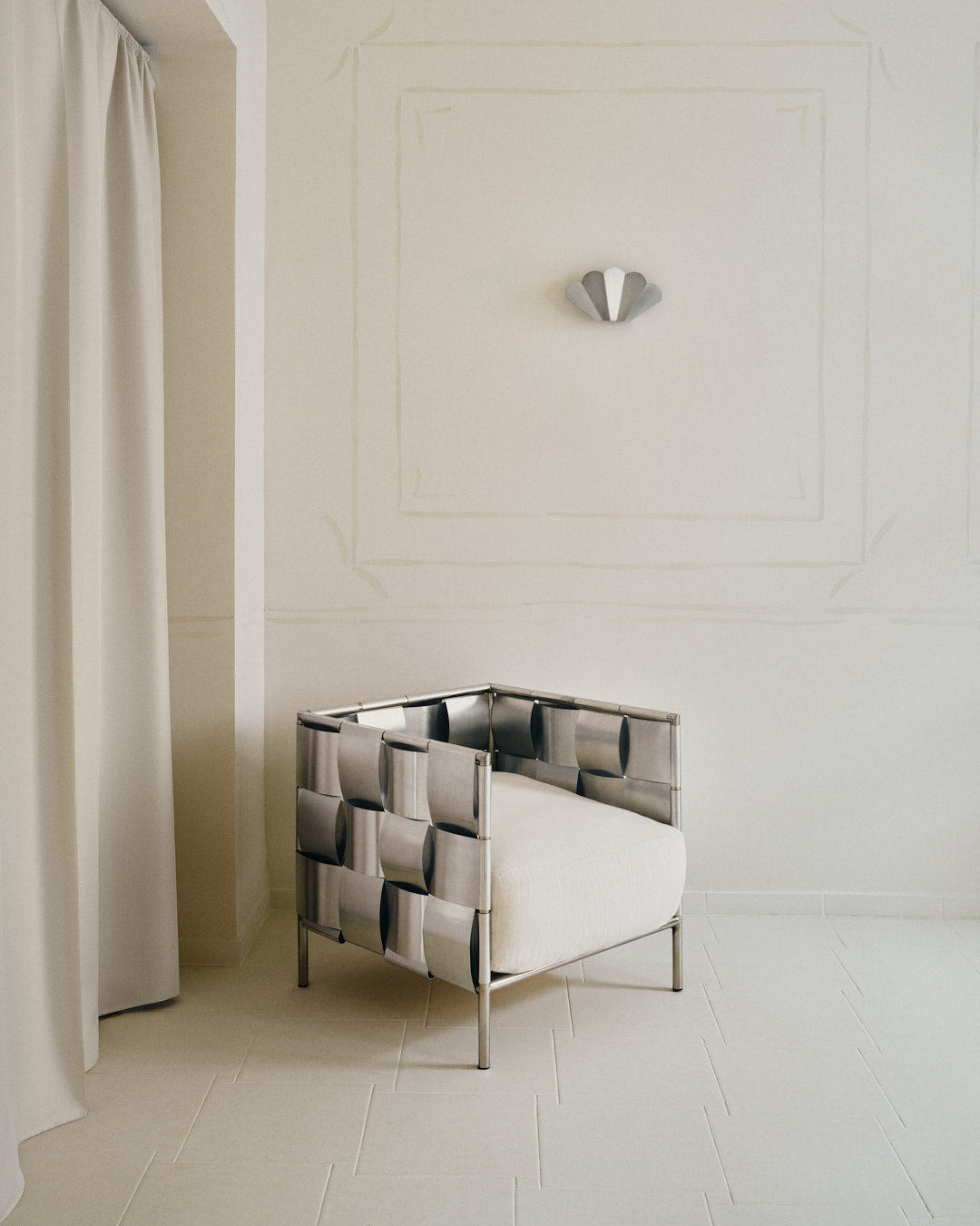 The Good Living&Co. "Braid" Armchair, featuring a metallic woven frame and white cushion, is placed in a minimal cream-toned room with ornate wall trim, sculptural sconce, and floor-to-ceiling curtains.