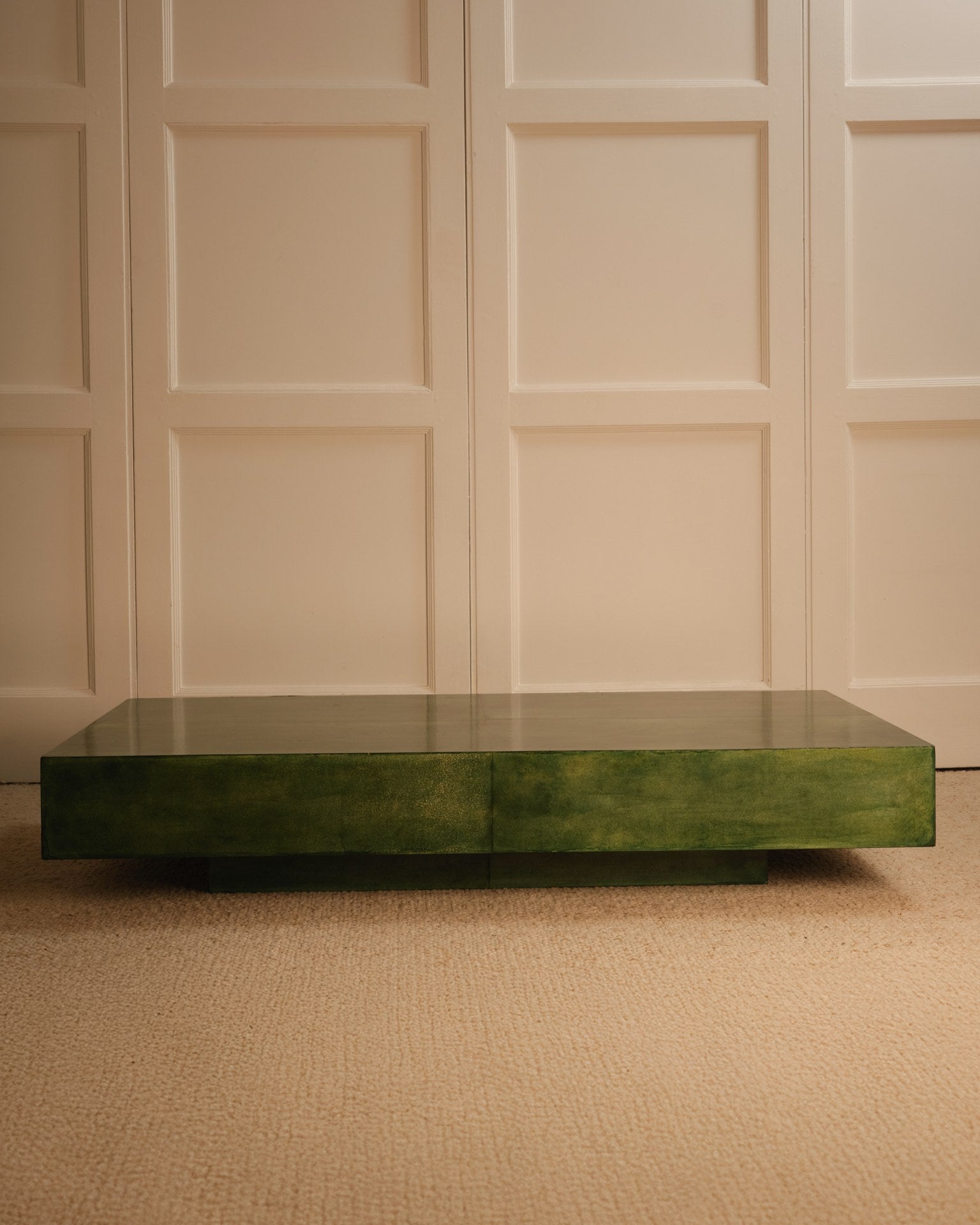 A Green parchment table by Galerie Vauvart, with a glossy rectangular surface, sits on a beige carpet before a cream-paneled wall in a softly lit, minimal space that echoes Aldo Tura’s signature style.