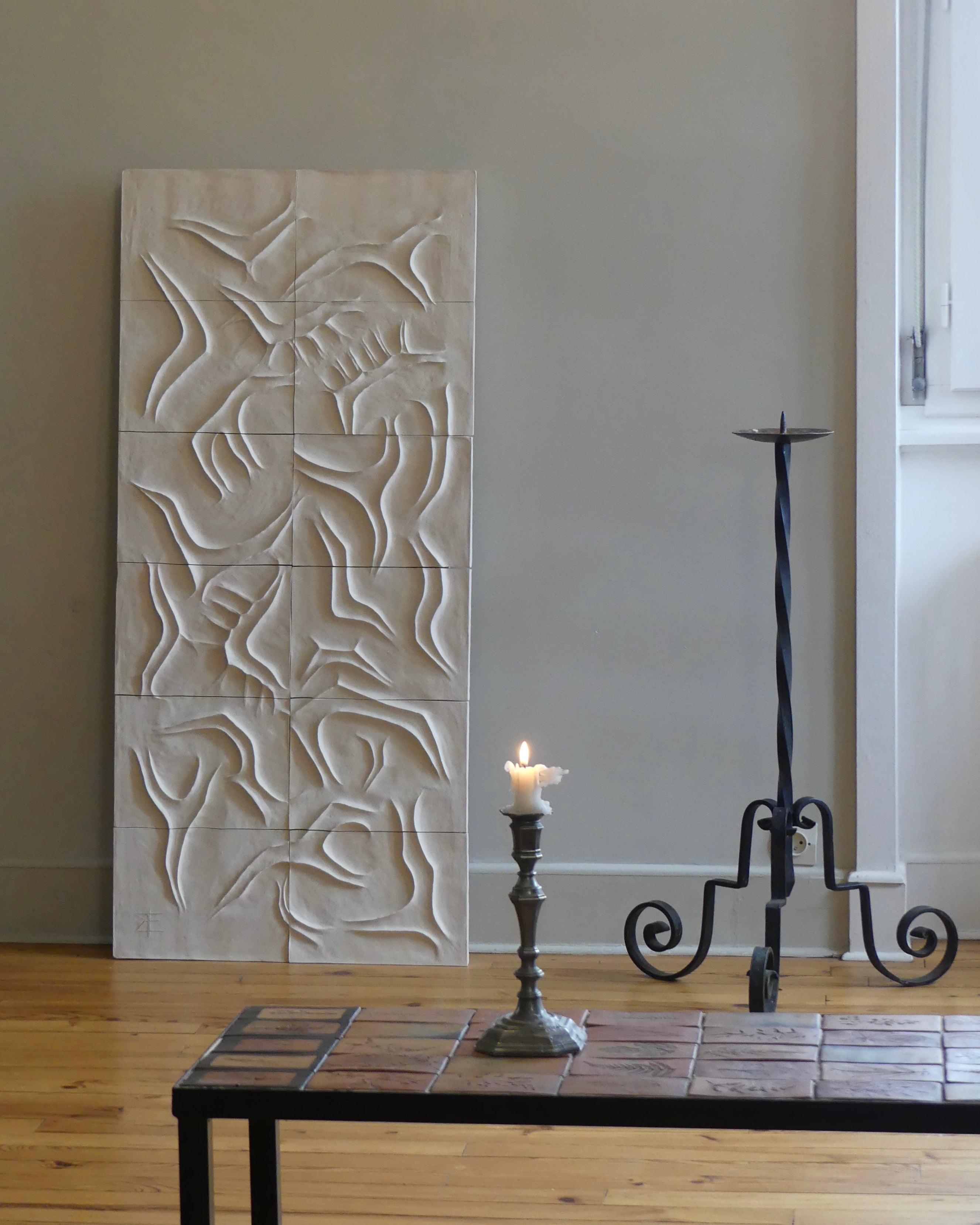 The Scala Dei Turchi Wall Sculpture by Franck Scala, with abstract white ceramic design, leans against a light gray wall beside a black metal candle holder and lit candle on a tiled table over a wooden floor.