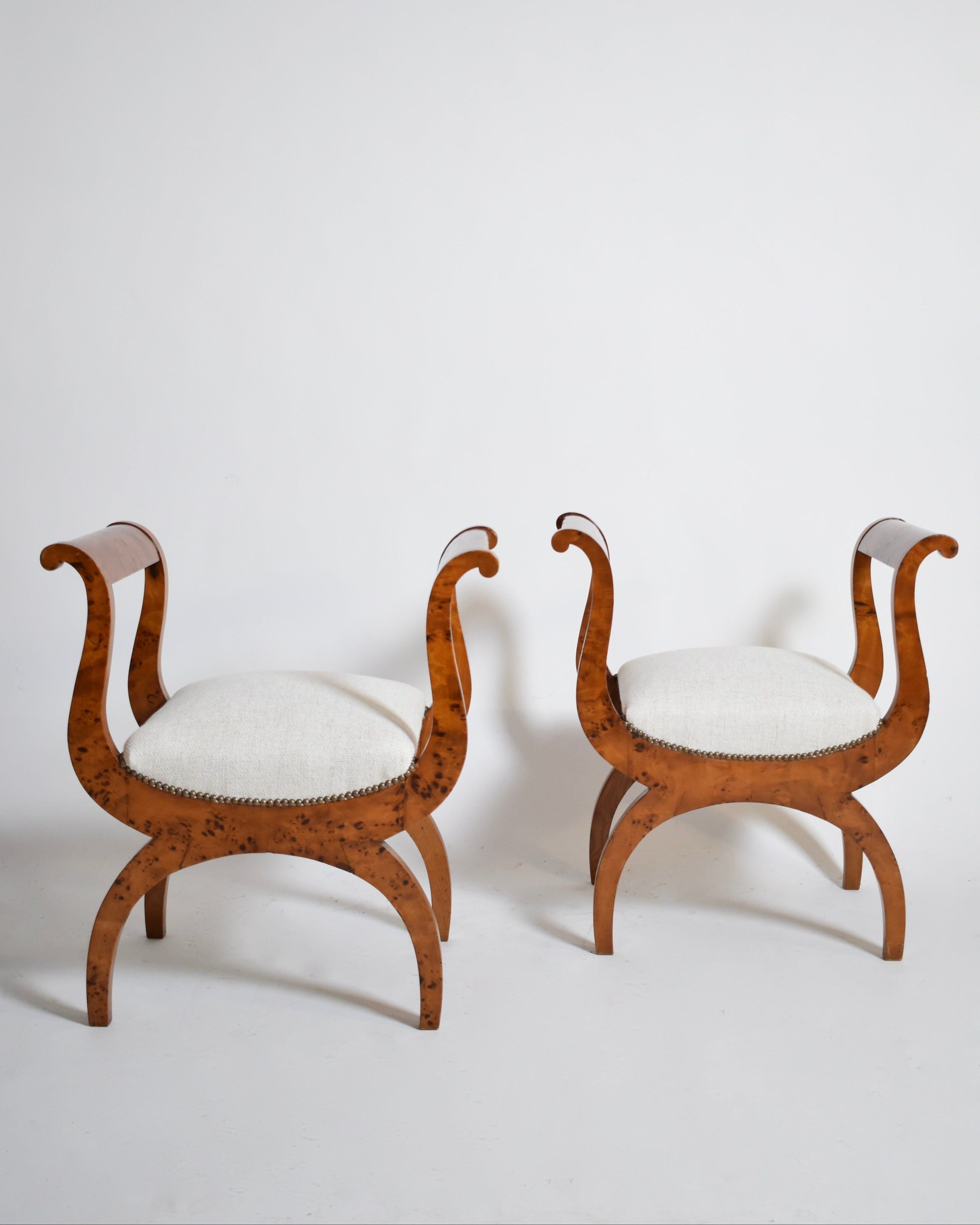 The Barracuda Interiors Pair of Walnut Curule Stools, XIXth, feature curved legs and arms with light upholstered seats. Their elegant symmetrical design and warm walnut tones stand out beautifully against a light background.