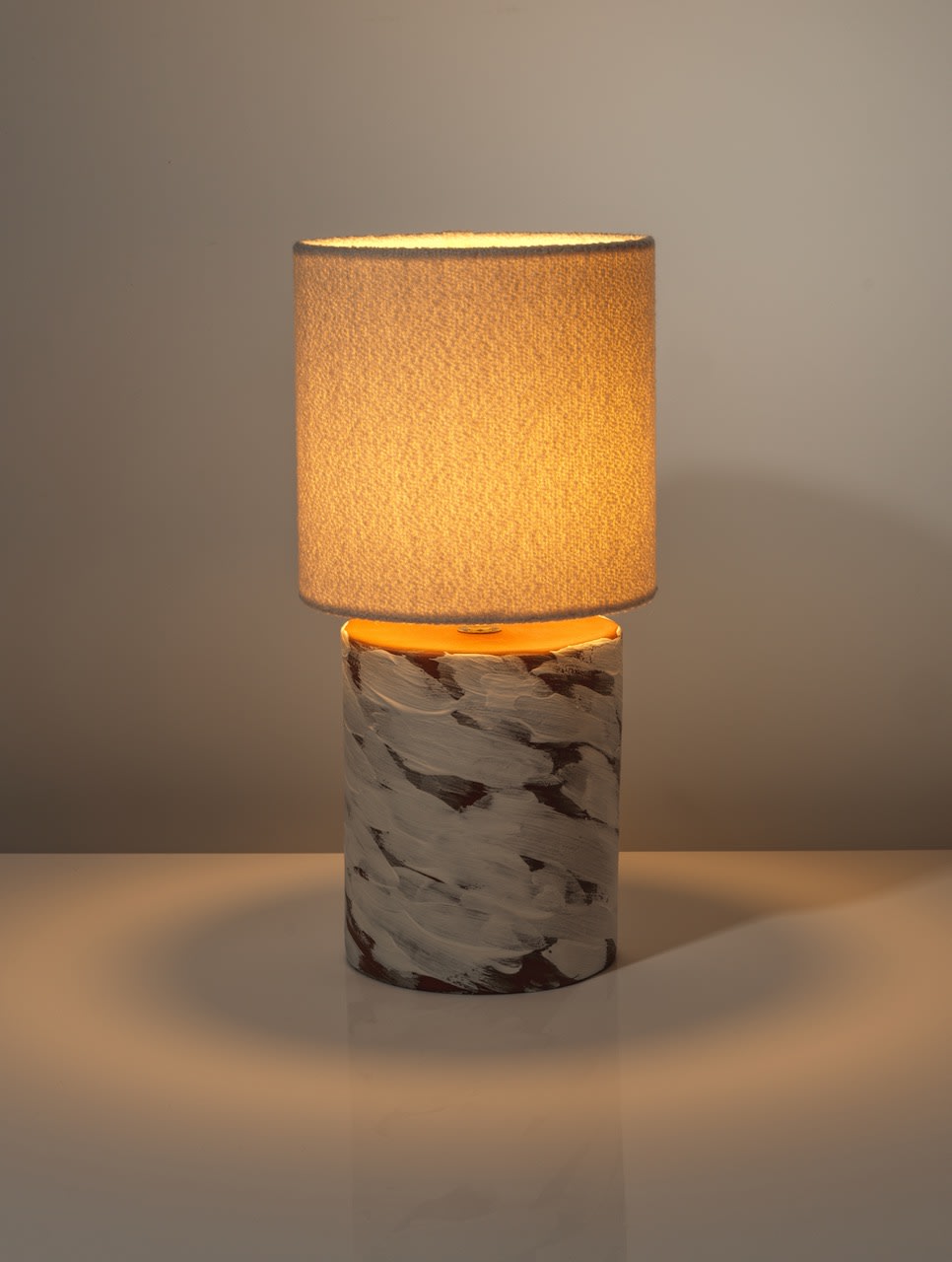 The White Wind Table Lamp by Katia Langeard features a modern ceramic base and a textured beige cylindrical shade, casting a warm glow on glossy white surfaces—perfect as a stylish bedside accent in neutral spaces.