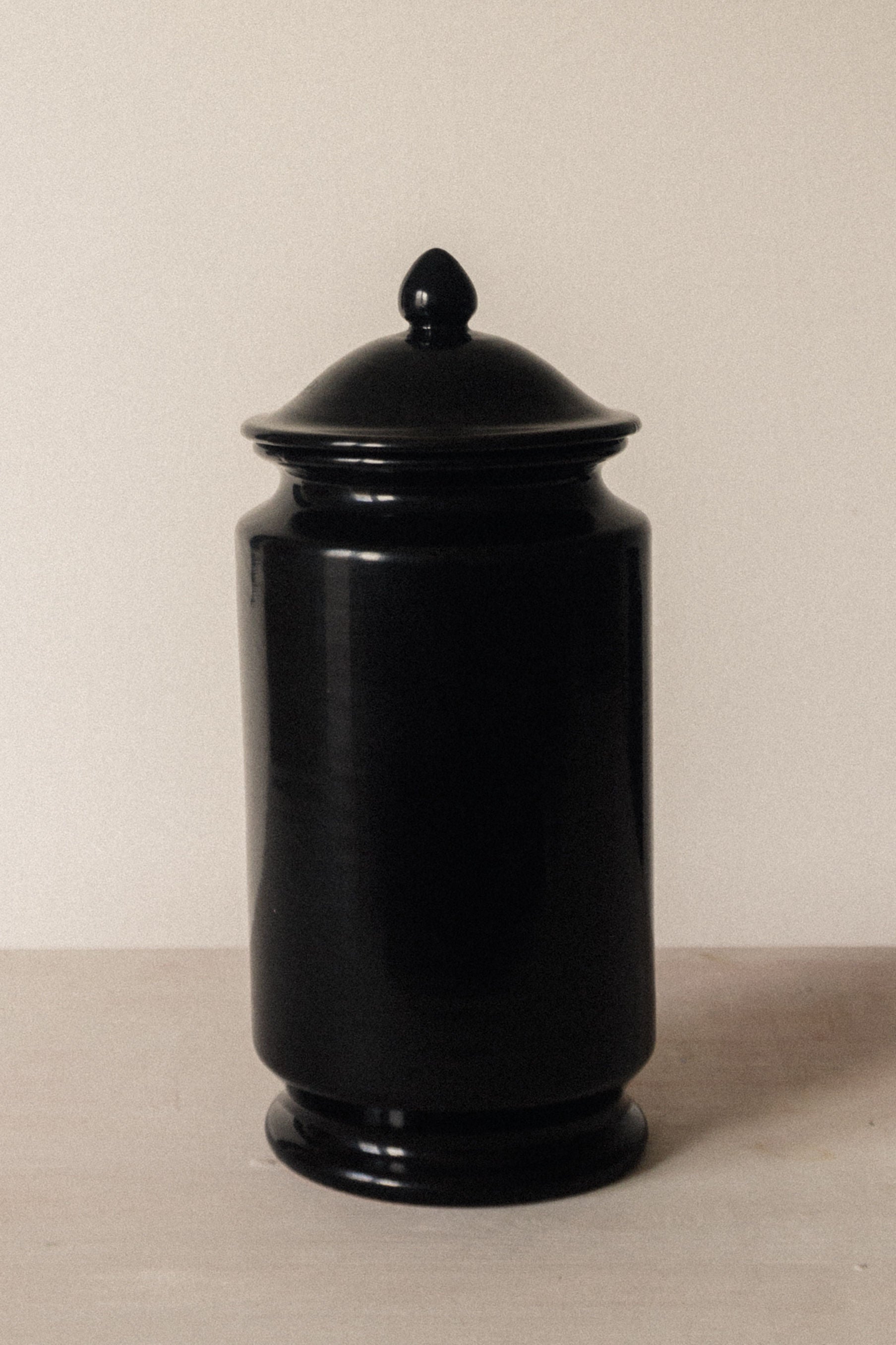 The Albarello - Minimalist Vase in Ceramic by COSEINCORSO, a glossy black jar with rounded lid and small knob, is displayed on a light surface against an off-white background.