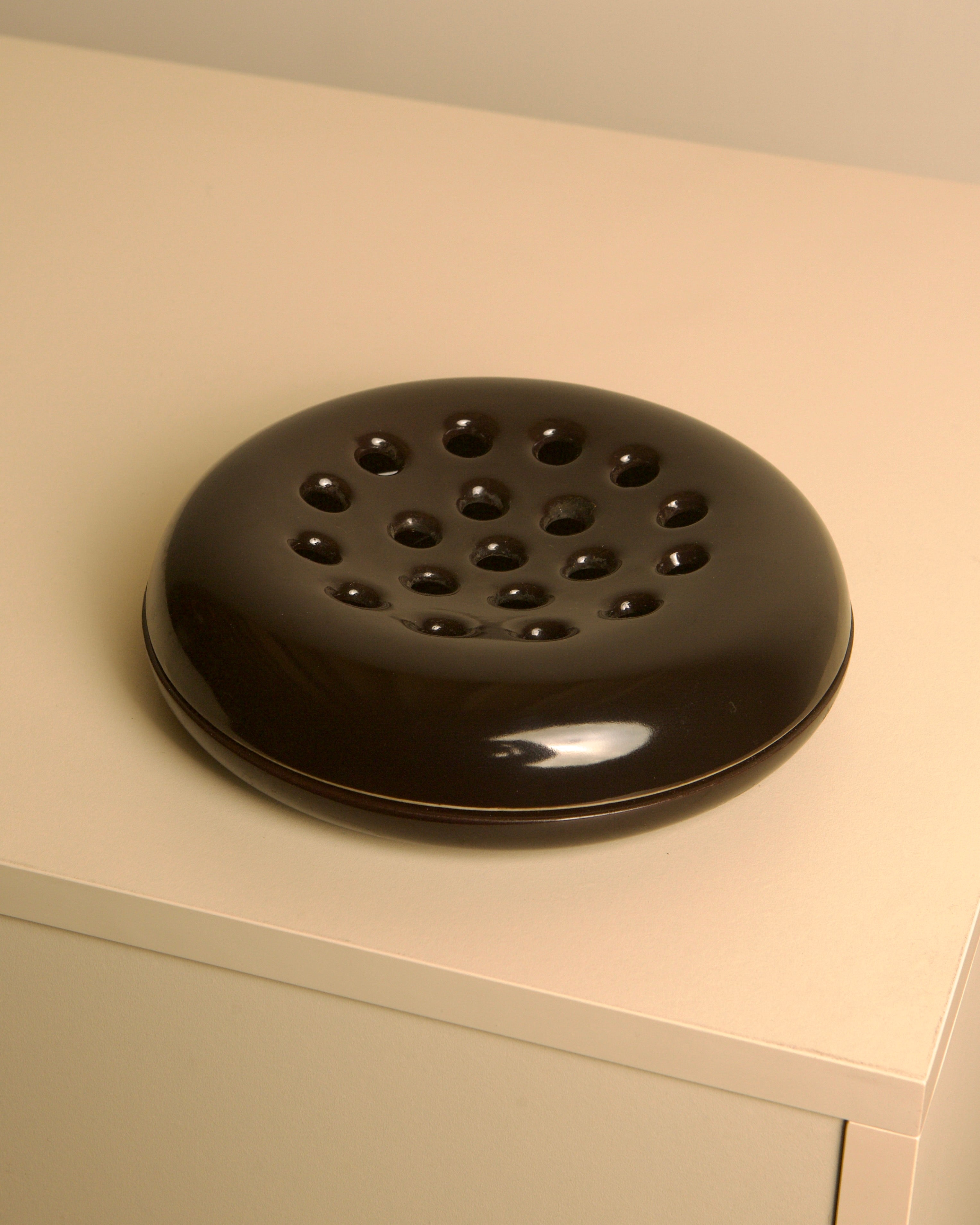 A Treaptyque Italian Brown Ceramic Ashtray by Angelo Mangiarotti for Brambilla, 1960s, featuring a round, glossy finish and multiple top holes, rests on a light beige surface.