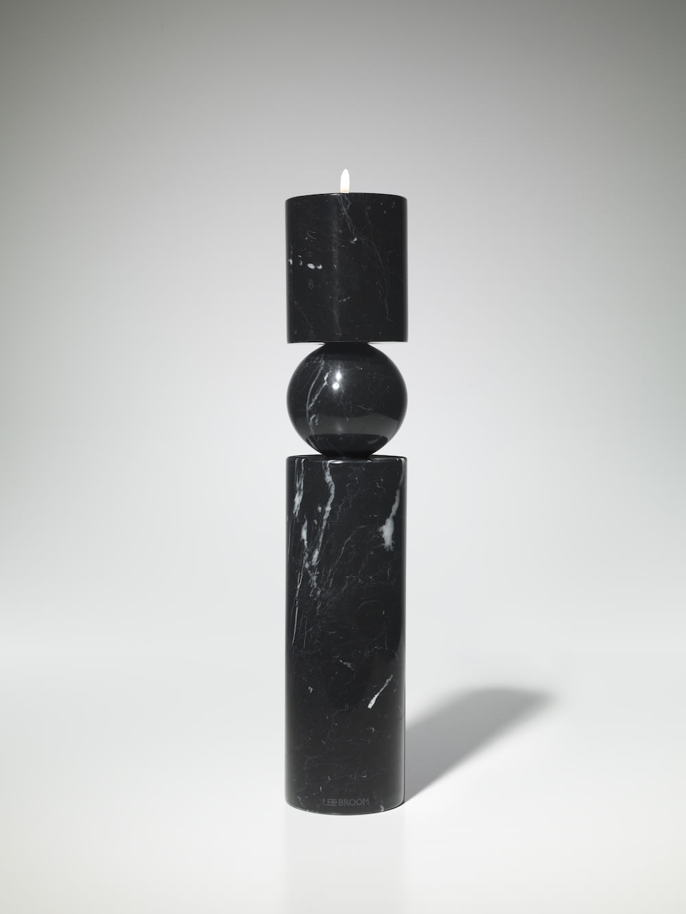 The Fulcrum Candlestick Black Marble by Lee Broom showcases three geometric forms—two cylinders and a sphere—crafted from black Marquina marble, elegantly holding a white candle against a simple, light background.