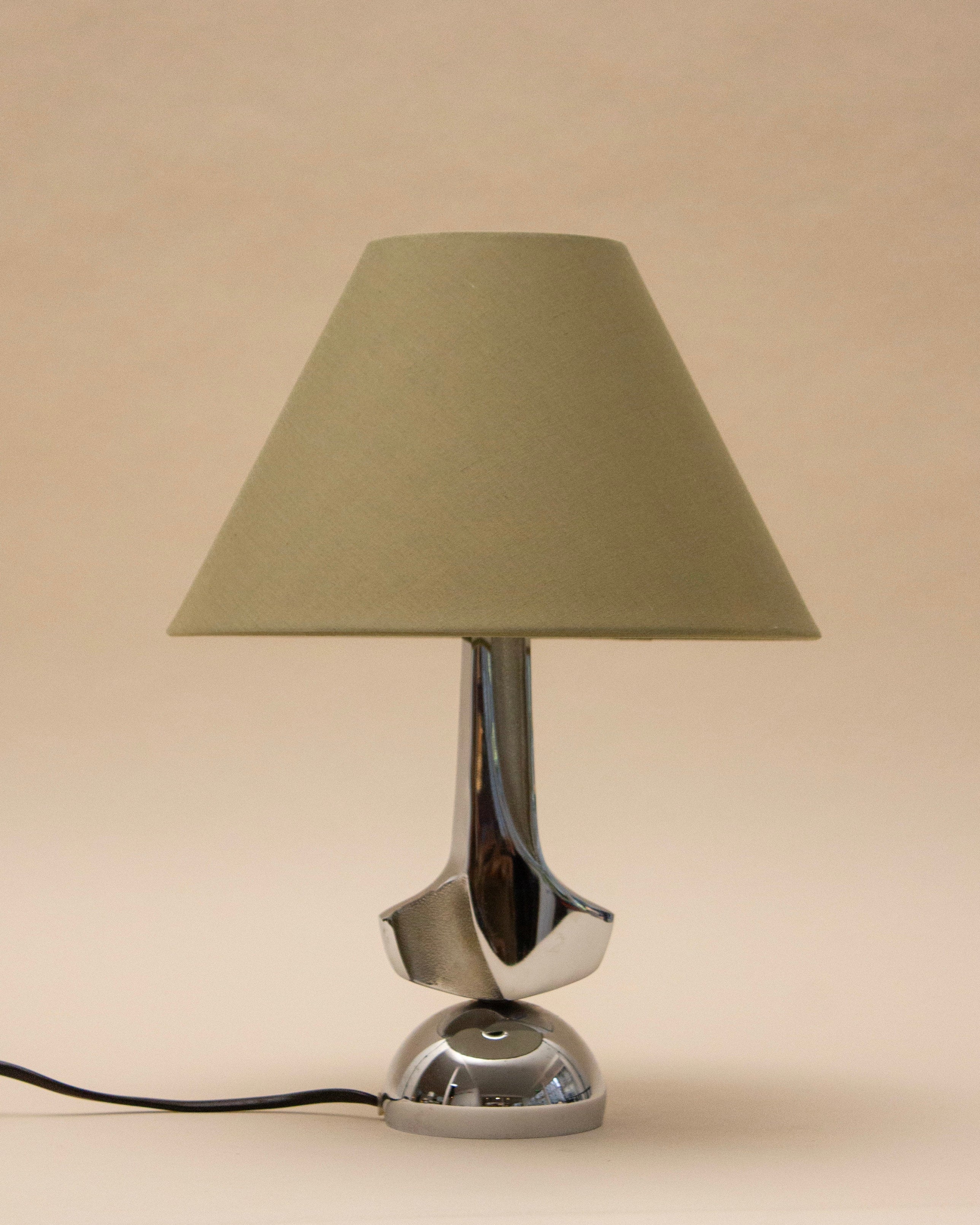 The SÒLER MELLILA TABLE LAMP features a modern chrome design with a beige conical shade and shiny silver base, set against a plain beige background. A black power cord extends from the base.
