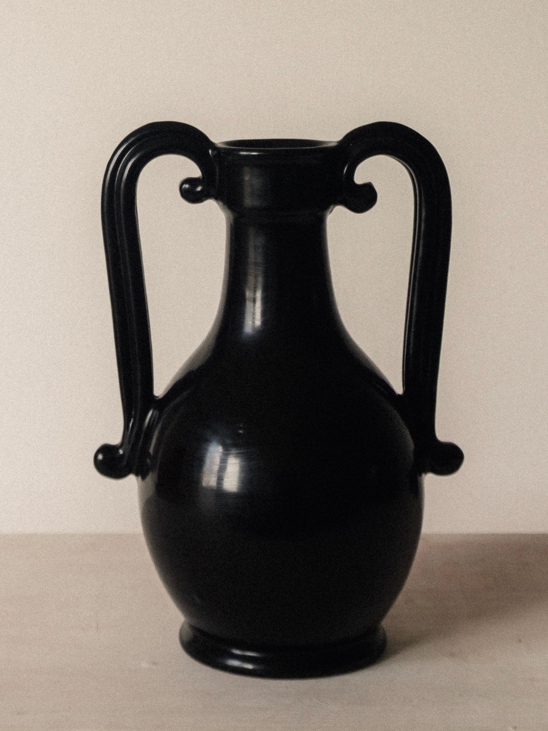 The Fiasca - Minimalist Vase in Ceramic by COSEINCORSO, a glossy black handmade Italian piece with two large curved handles, stands on a light surface against an off-white background.