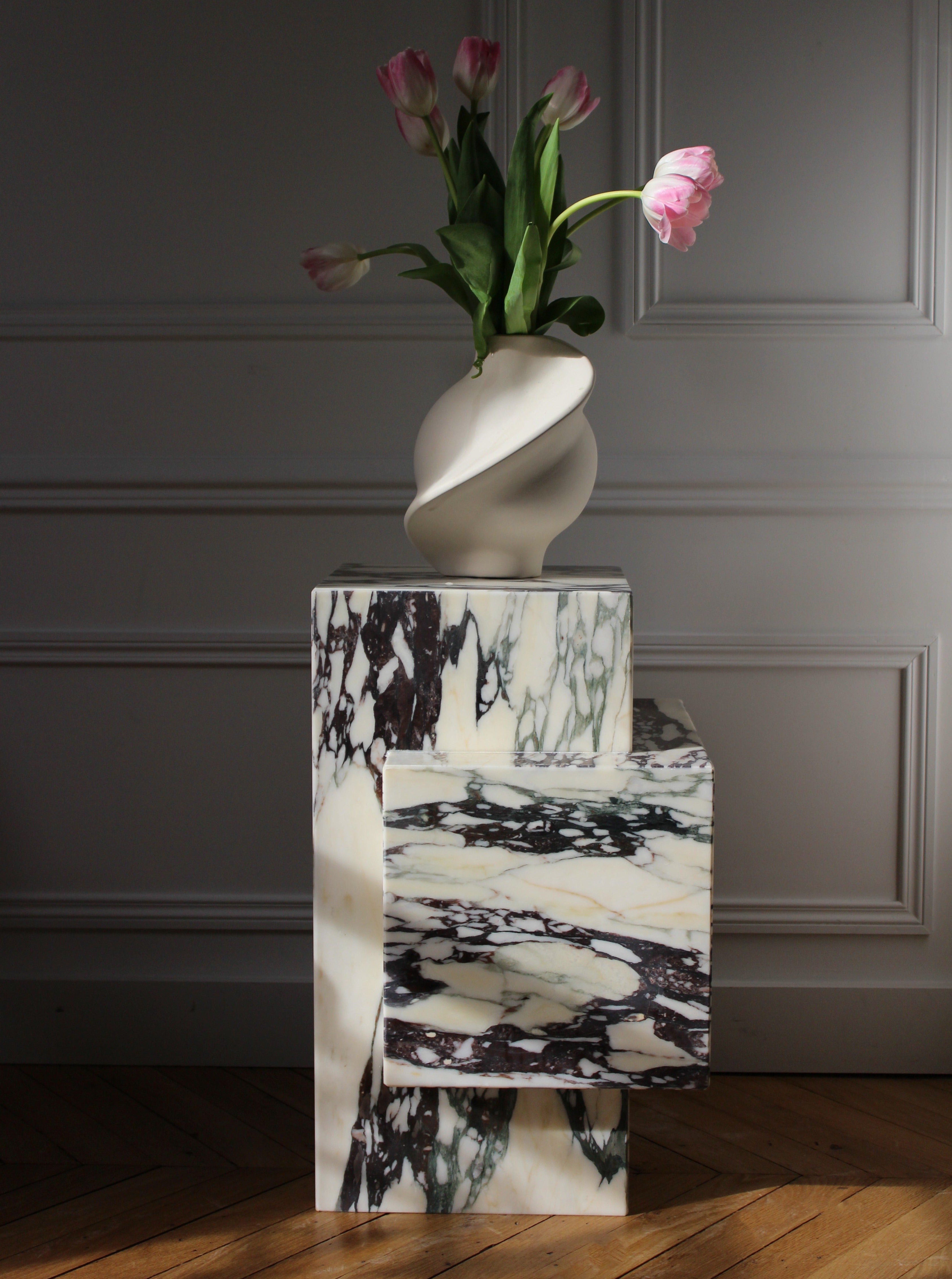 The Ella Calacatta Viola Marble Table from Marbera, featuring black and white patterns, supports a sculptural vase with pink tulips. This minimalist design accents a room with elegant paneling and wooden floors, where sunlight streams in from the side.