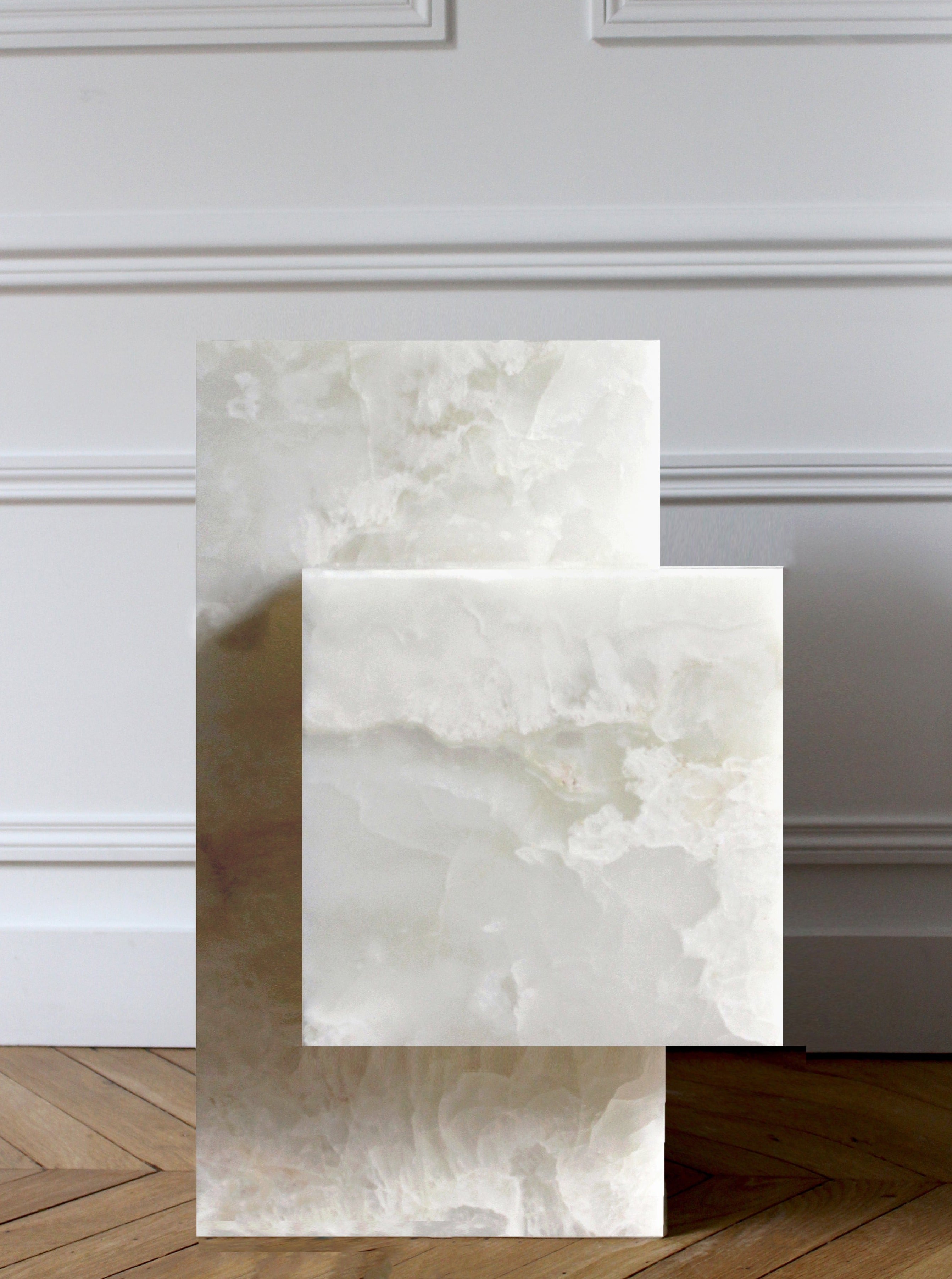 The Ella White Onyx - Marble Coffee Table by Marbera, featuring two stacked rectangular slabs with a marble texture, stands on a chevron-patterned wooden floor against a white paneled wall. Its translucent appearance in elegant white and gray shades enhances the luxurious interior.