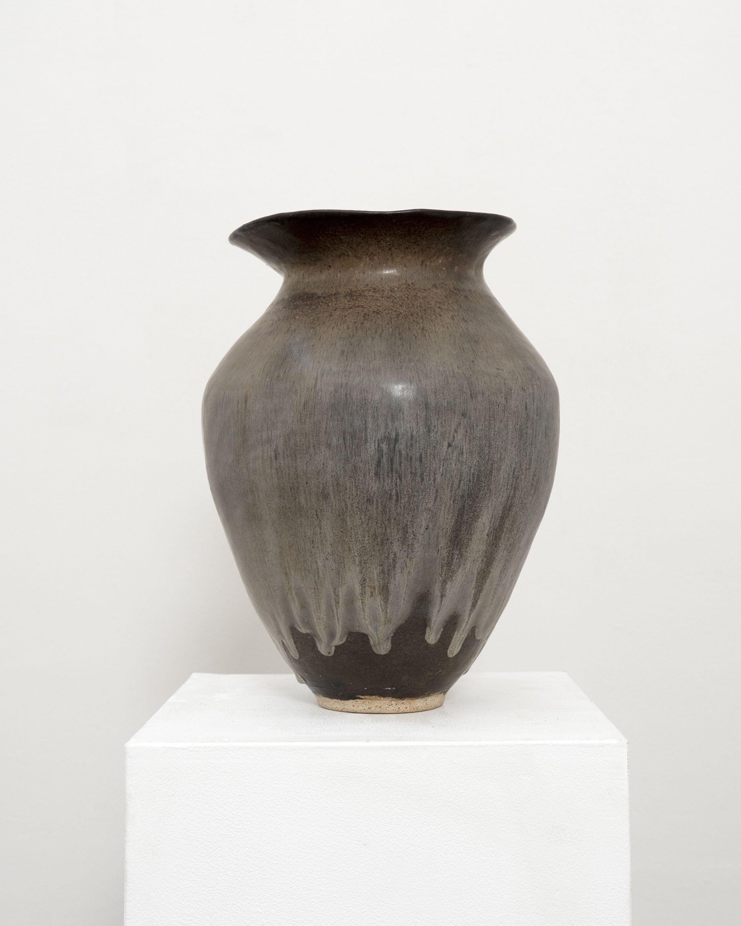 The Hand Built Studio Pottery Vase by Tom Bogle features a wide, flared mouth, rounded body, and brown-gray gradient glaze. This stoneware piece is displayed on a white pedestal against a light background.