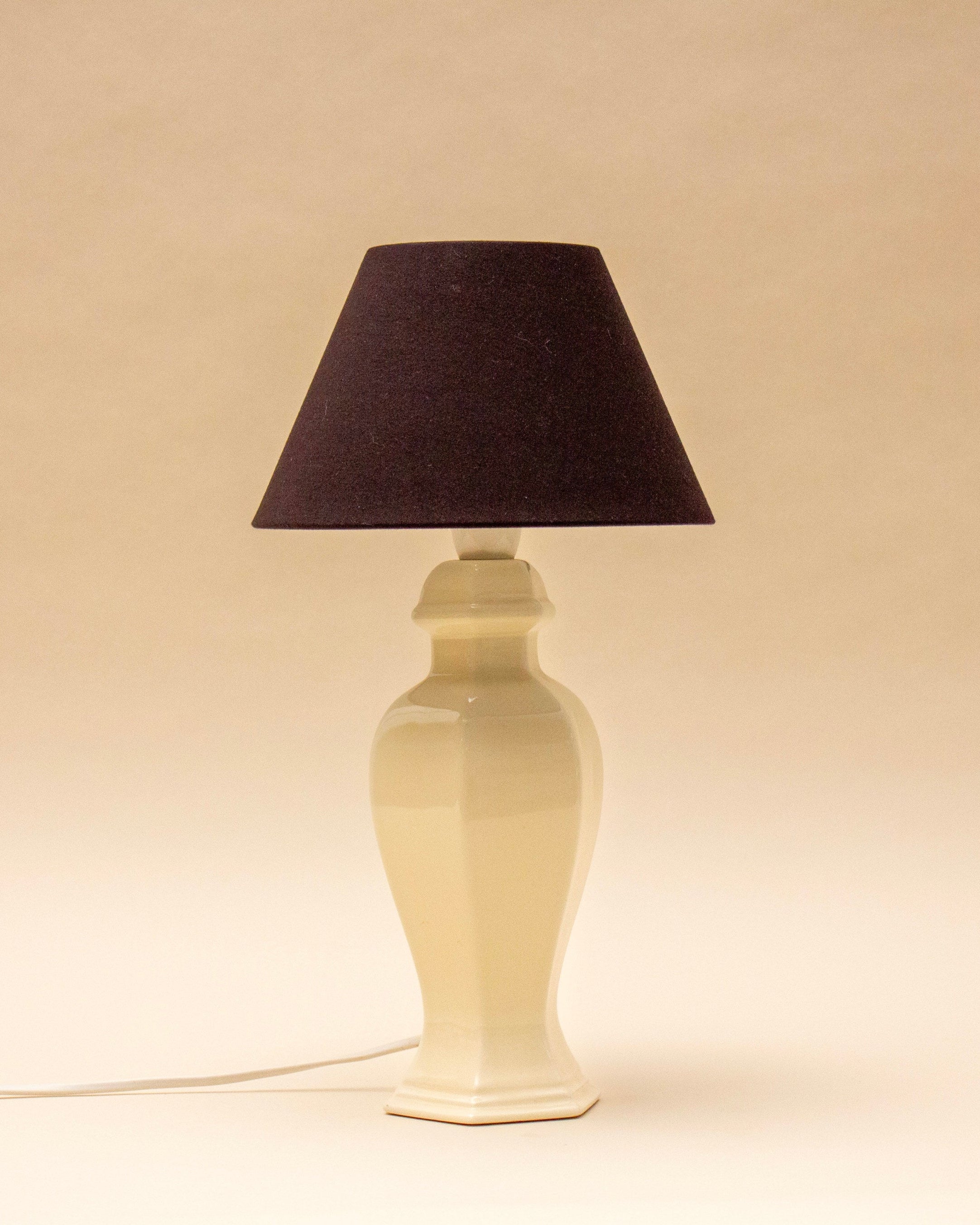 The SÒLER ALICANTE TABLE LAMP, featuring a beige ceramic base and black lampshade, stands on a light surface with its vintage-style cord visible against a plain beige background.