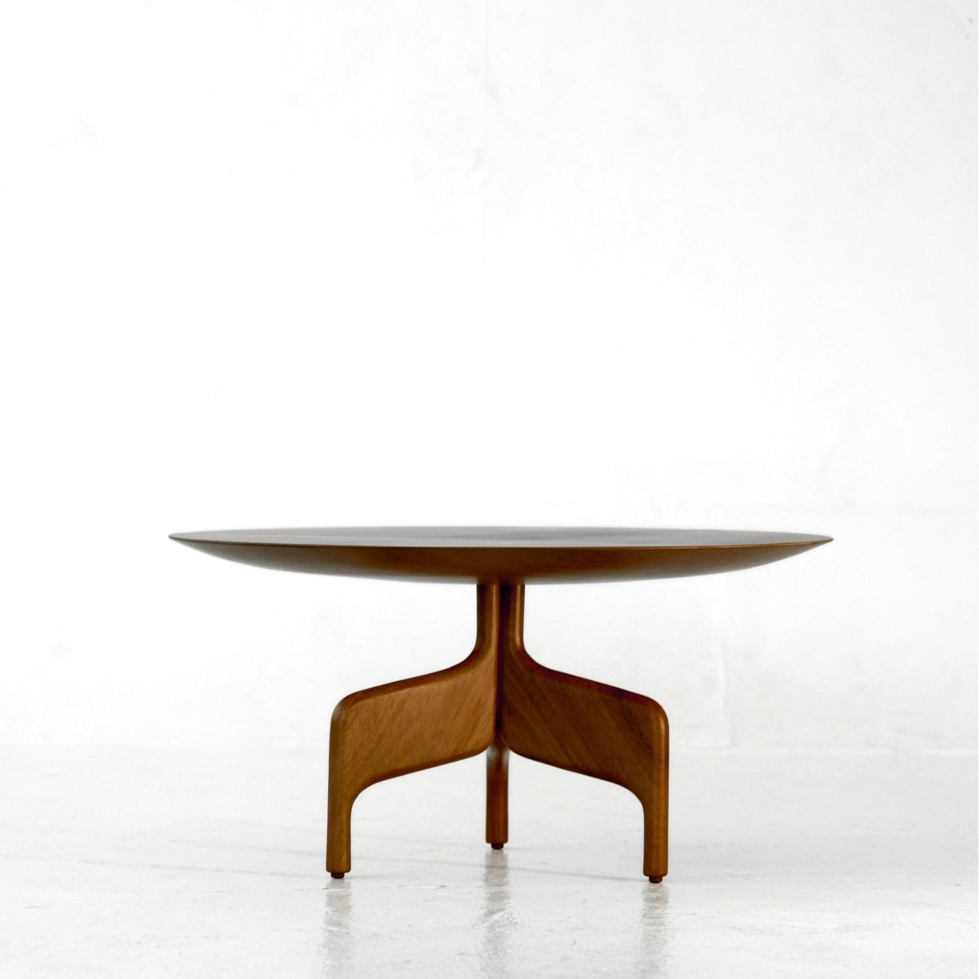 The "Alaia" Table by Luciano Costa Estúdio is a round wooden piece from the Nômade collection, featuring a smooth top and distinctive three-legged solid lyptus wood base, shown on a plain white background.