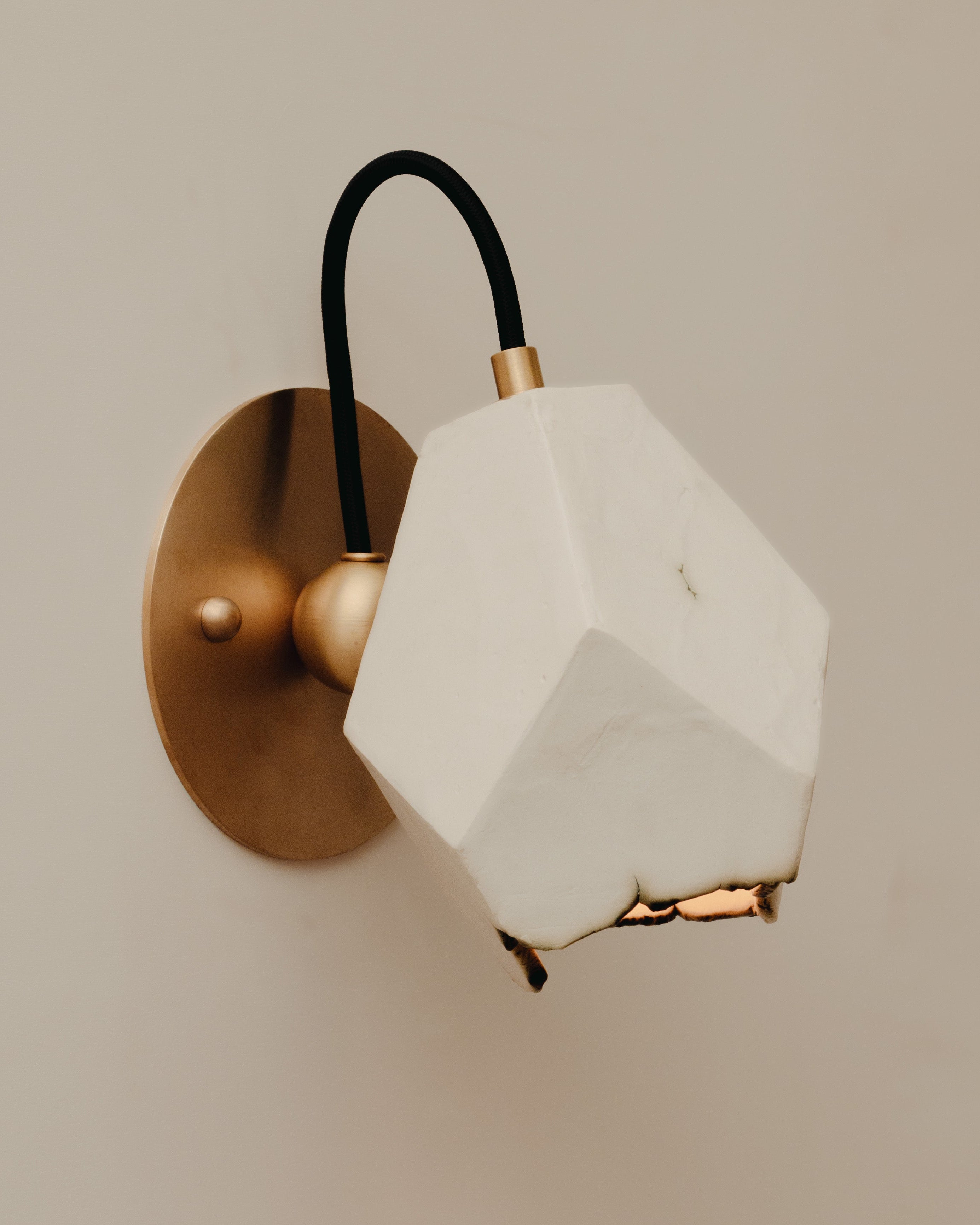 The Relic Spotlight Sconce by JS/WRKSHP features a geometric white porcelain shade and brass base, connected by a black cord, for a contemporary look. It is mounted on a beige wall.