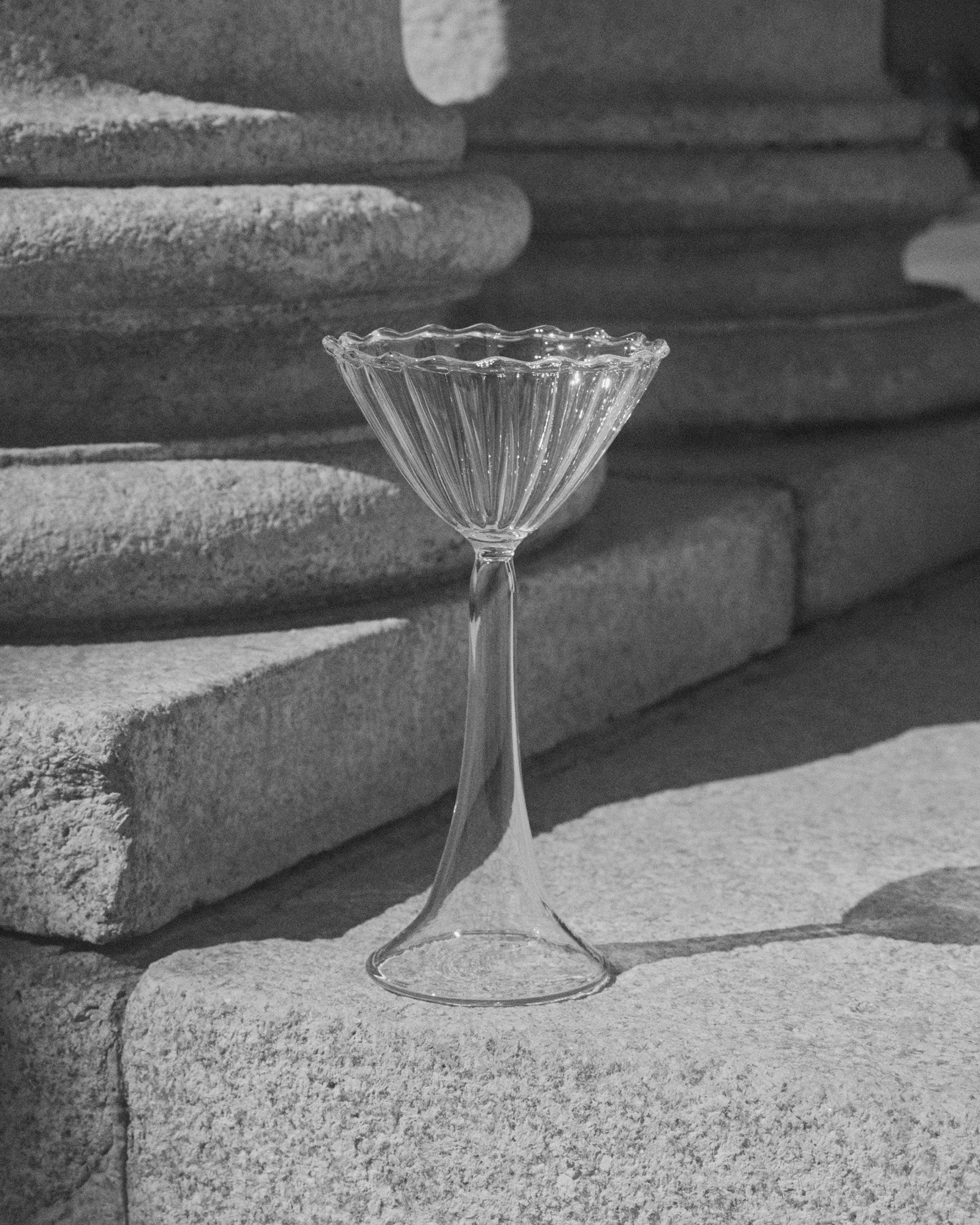 The Campanula — Clear Cocktail Glass by Agustina Bottoni stands on stone steps in sunlight, set against textured columns. A black-and-white photo highlights the elegant handmade borosilicate glass artistry from Italy.