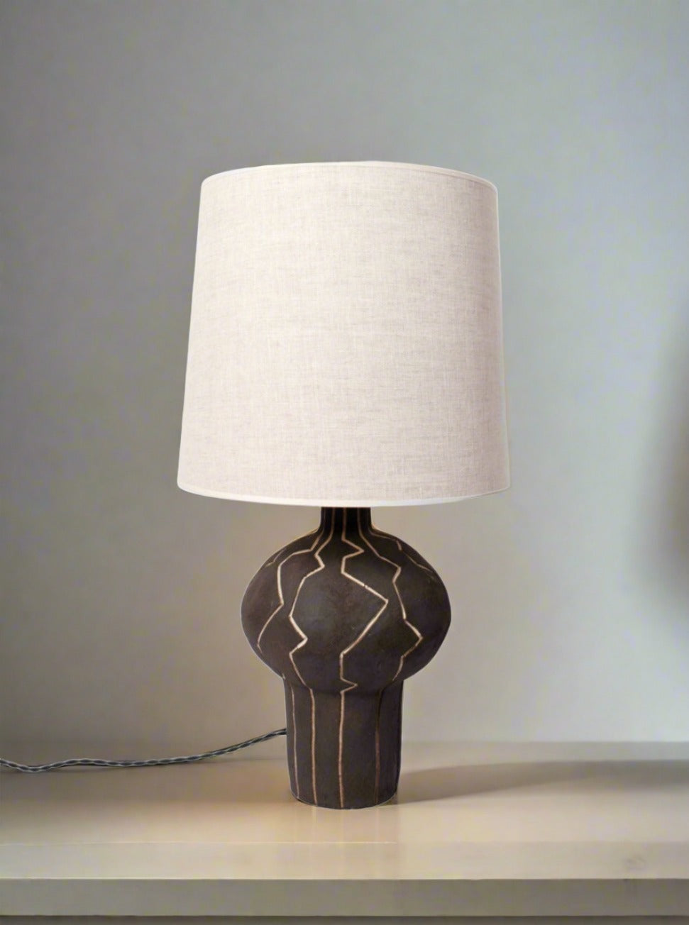 The "Lanzarote" Lamp by Barracuda Interiors features a black ceramic base with geometric lines and a large off-white fabric shade, providing customizable lighting on light surfaces in neutral spaces.