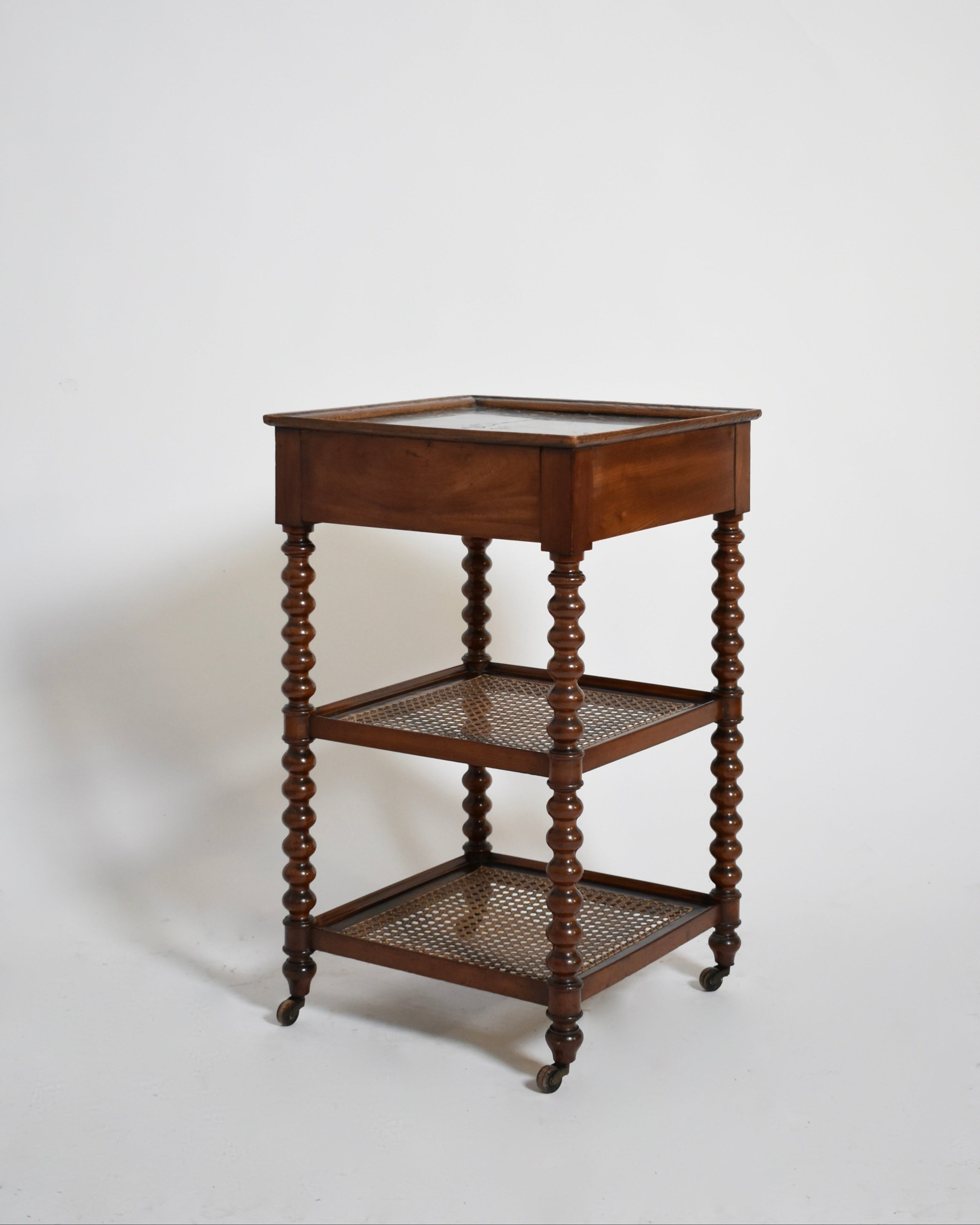 The Barracuda Interiors Serving Table Two Trays, XIXth c., features cane trays, a glass-enclosed top shelf, turned spindle legs, and small wheels—an elegant vintage piece shown against a white background.