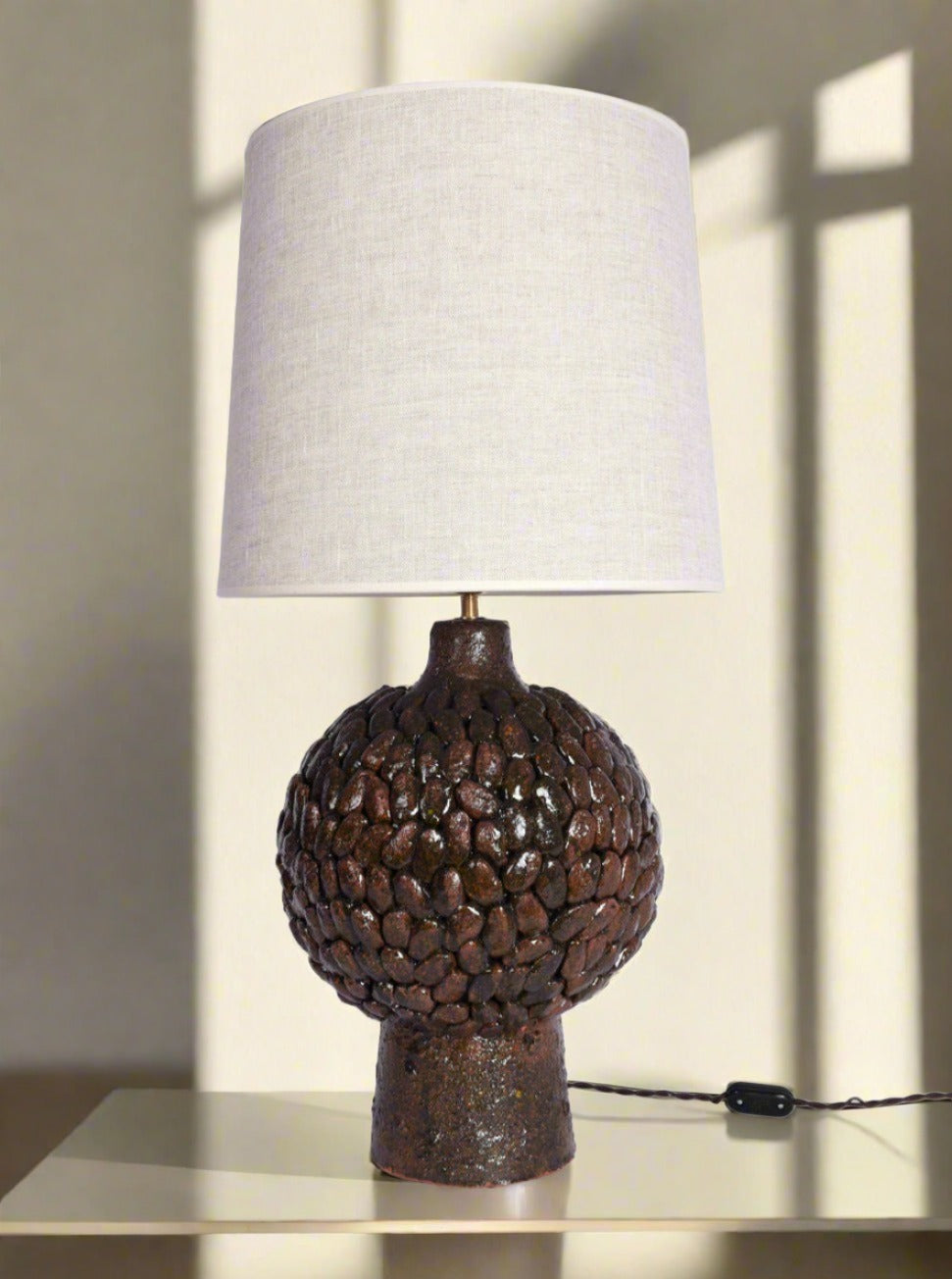 The "Melides" Brown Glazed Terracota Lamp by Barracuda Interiors features a round, textured base resembling clustered stones and a plain white lampshade, shown on a light surface against a wall with window-shadow patterns. Barracuda Edition.
