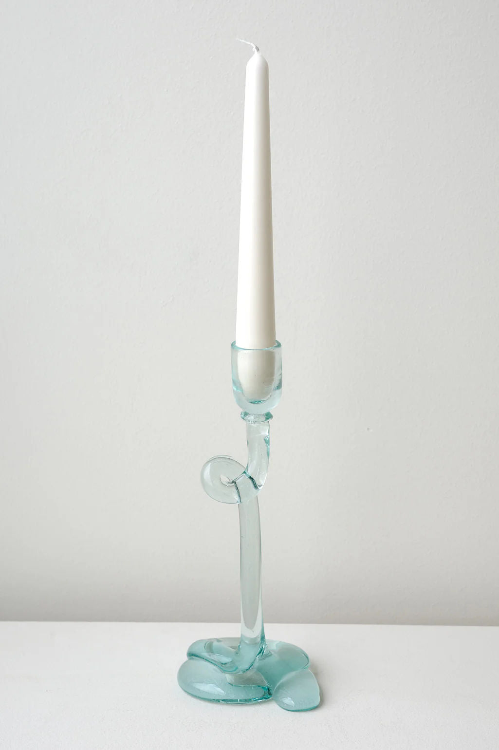 A white taper candle sits in the Thaw - Recycled Glass Candleholder in Clear by Completedworks, featuring an abstract wavy design, placed on a white surface with a plain light gray background.