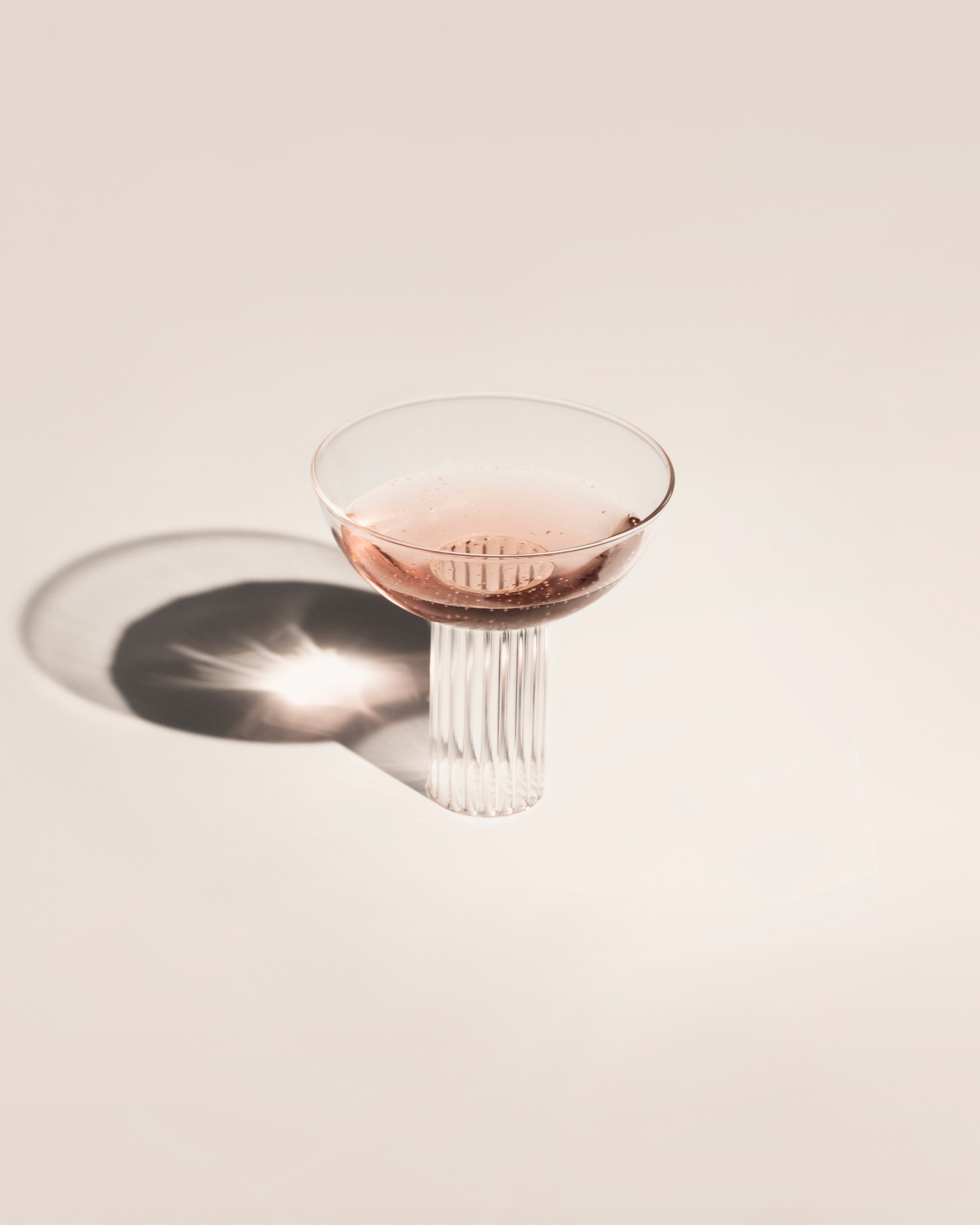 Pink-tinted coupe glass with ribbed stem casting elegant shadow