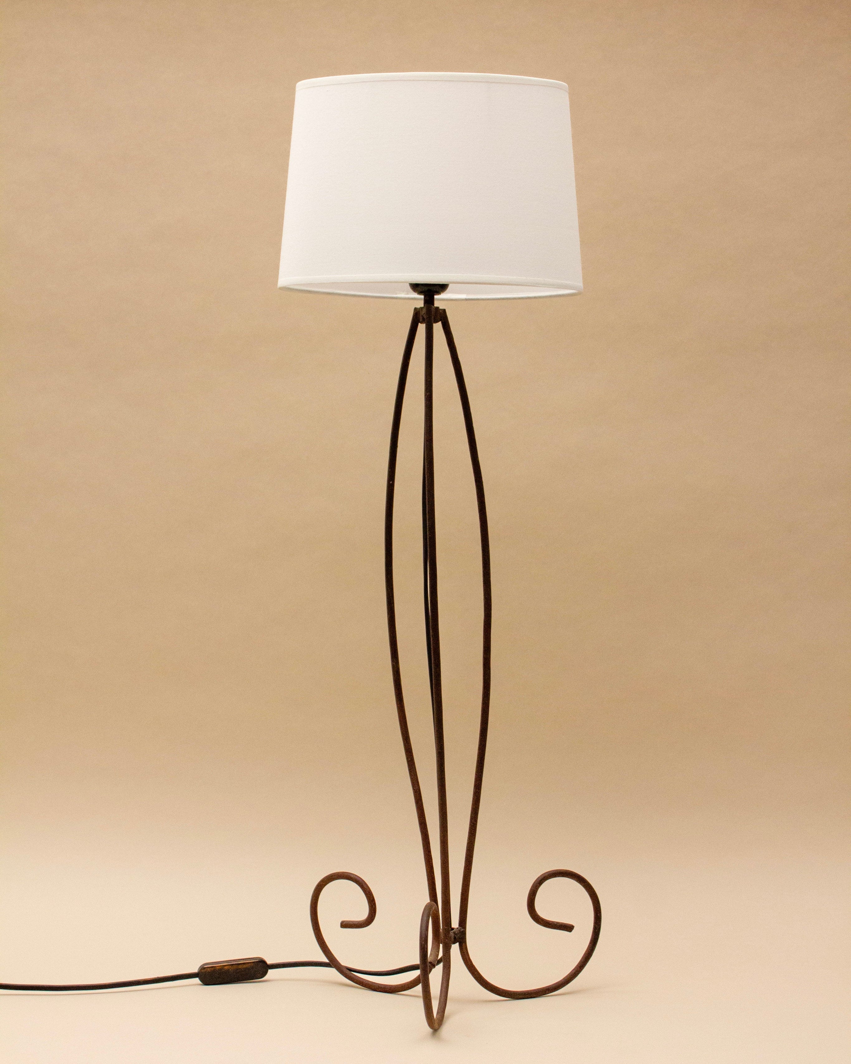 The SÒLER SANTA CRUZ DE TENERIF LAMP is a tall floor lamp with a vintage shade and slender brown metal base featuring elegant curved scroll designs, shown here on a plain beige background.