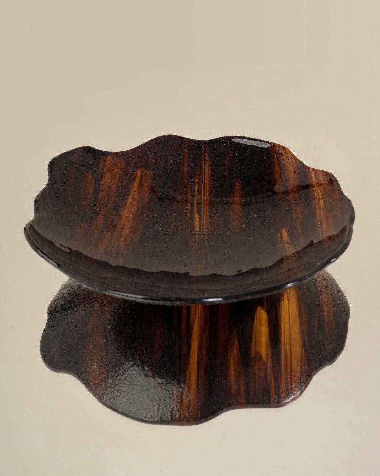 A dark, wooden, wavy-edged Plate Carey Fig by Los Objetos Decorativos displayed on a reflective surface, creating a clear shadow below it. The plate's rich brown tones have swirling patterns characteristic of artisanal manufacturing.